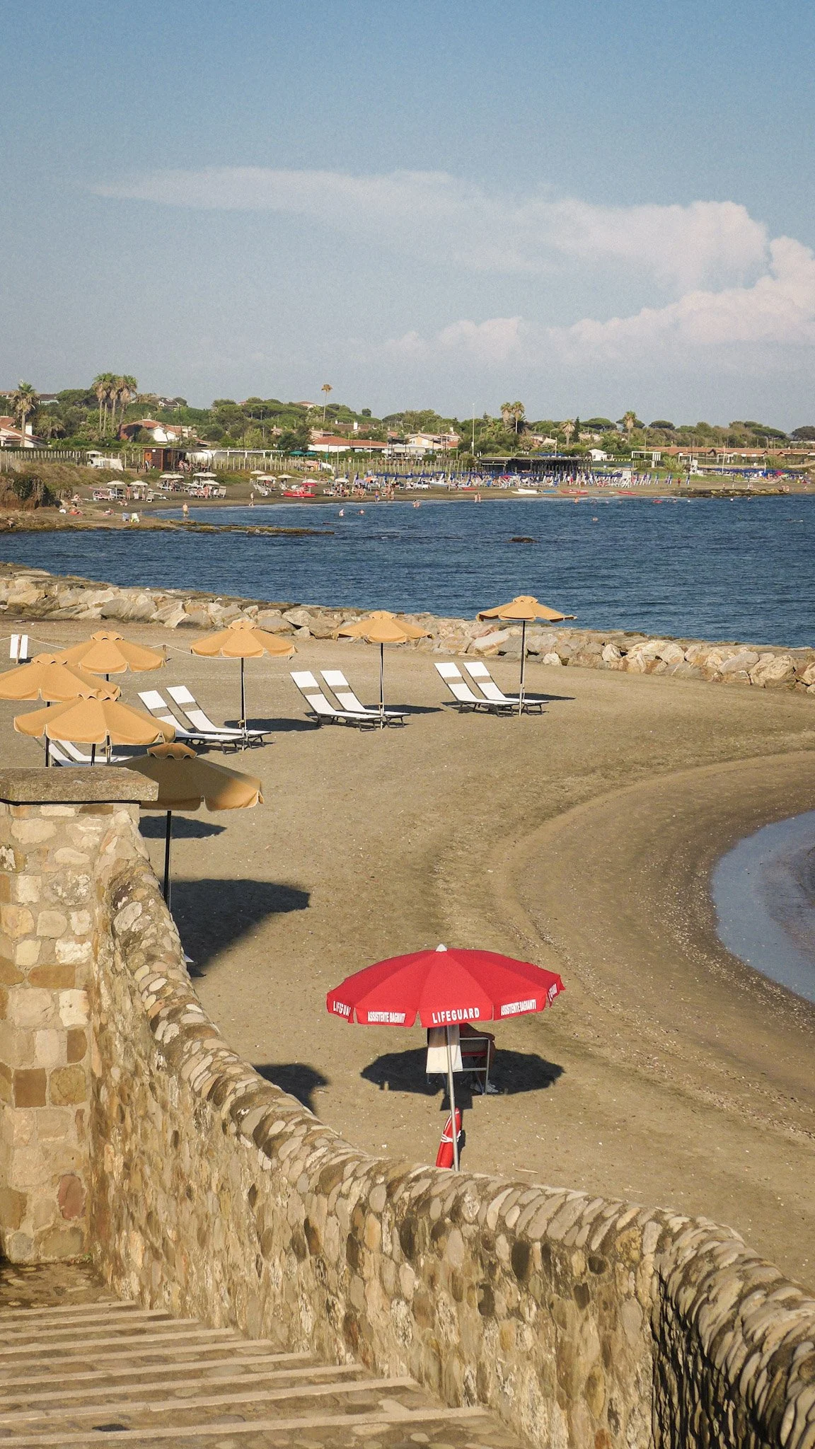 View of a beach with orange umbrellas, a lifeguard umbrella, and sandy pathway, overlooking the water with a rocky shoreline, beachgoers, and houses in the background under a partly cloudy sky.