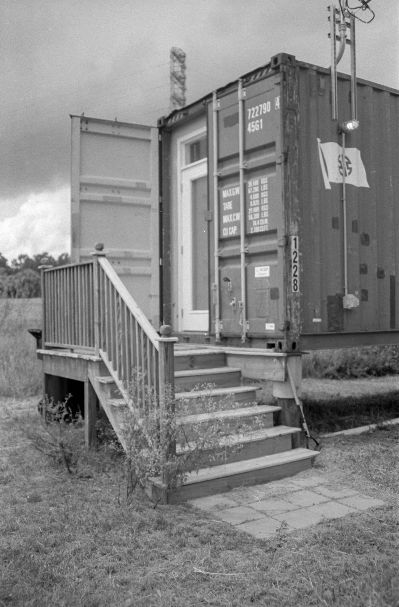 Black and white photo of a house with a small set of stairs leading up to a porch, which is surrounded by a railing. There are bushes and grass in the yard, with a sidewalk in front. The house has a door with a window and a small window on either sid