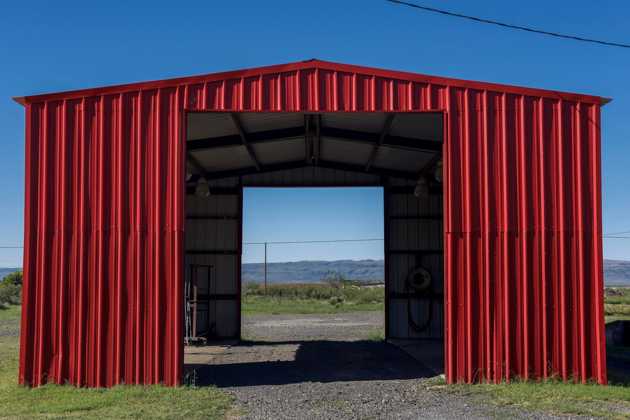Red metal barn with open front, opening to a landscape of green grass, gravel, and mountains under a clear blue sky.