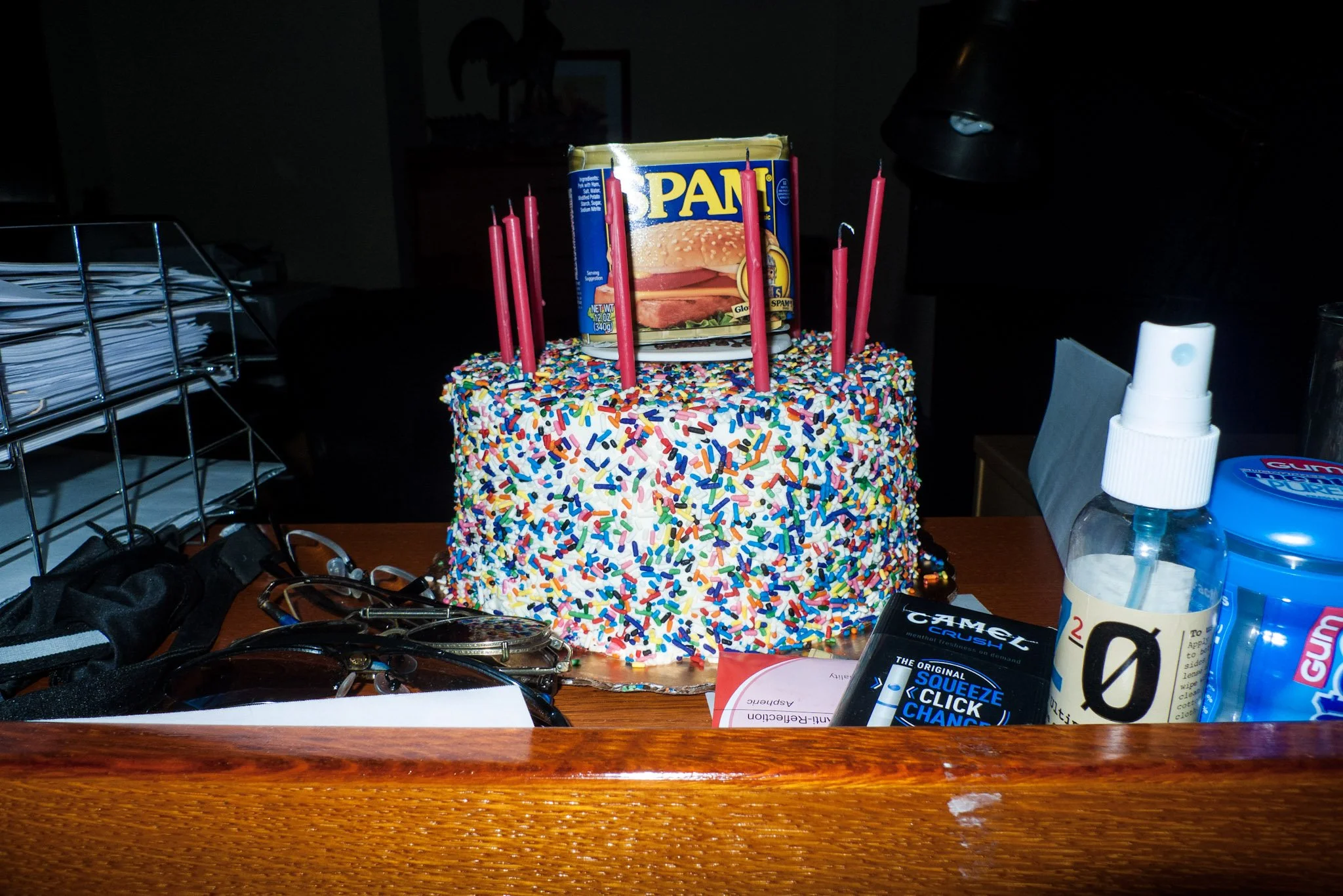 A birthday cake covered in colorful sprinkles with a can of Spam on top, surrounded by pink candles in a dimly lit room.