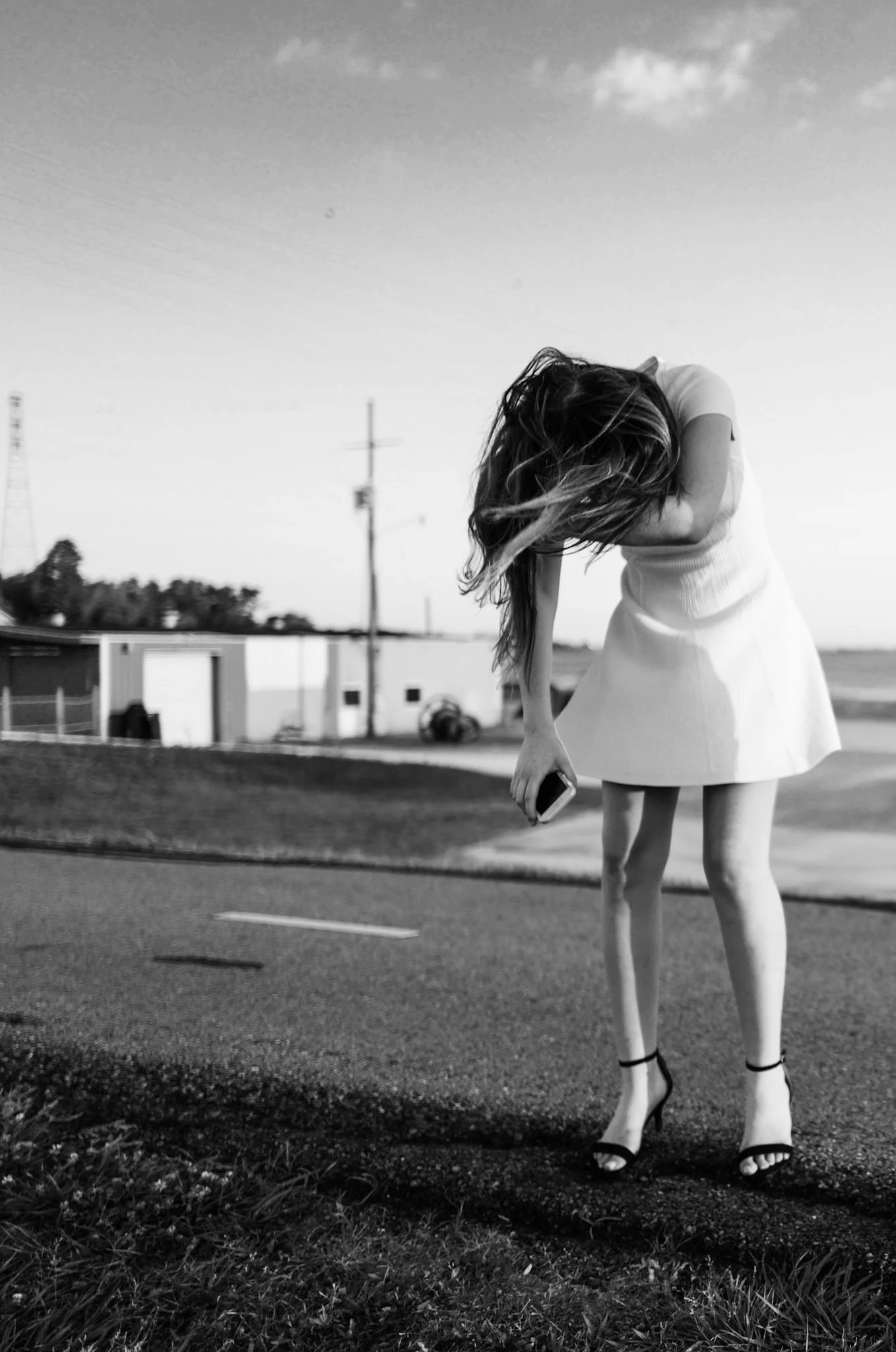 A woman with long hair, dressed in a light-colored dress and high heels, standing outdoors on a patch of grass and asphalt, holding a phone, with her head bowed and hair covering her face. In the background, utility poles, power lines, and buildings 