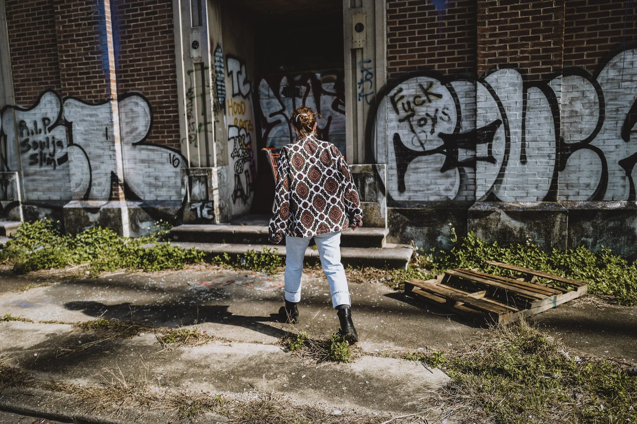A woman with her hair in a bun and wearing a patterned jacket, white pants, and black boots is standing in front of a graffiti-covered brick wall, facing away from the camera.