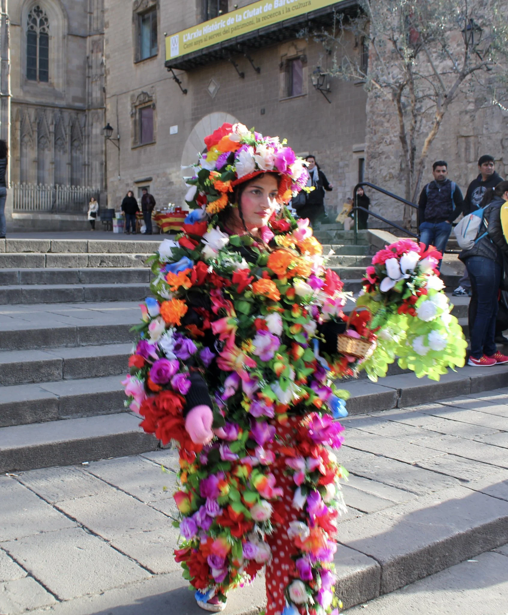 A person dressed in a costume made of colorful artificial flowers, wearing a large floral hat, walking down stone steps in an urban setting with a historical building and a small group of onlookers in the background.