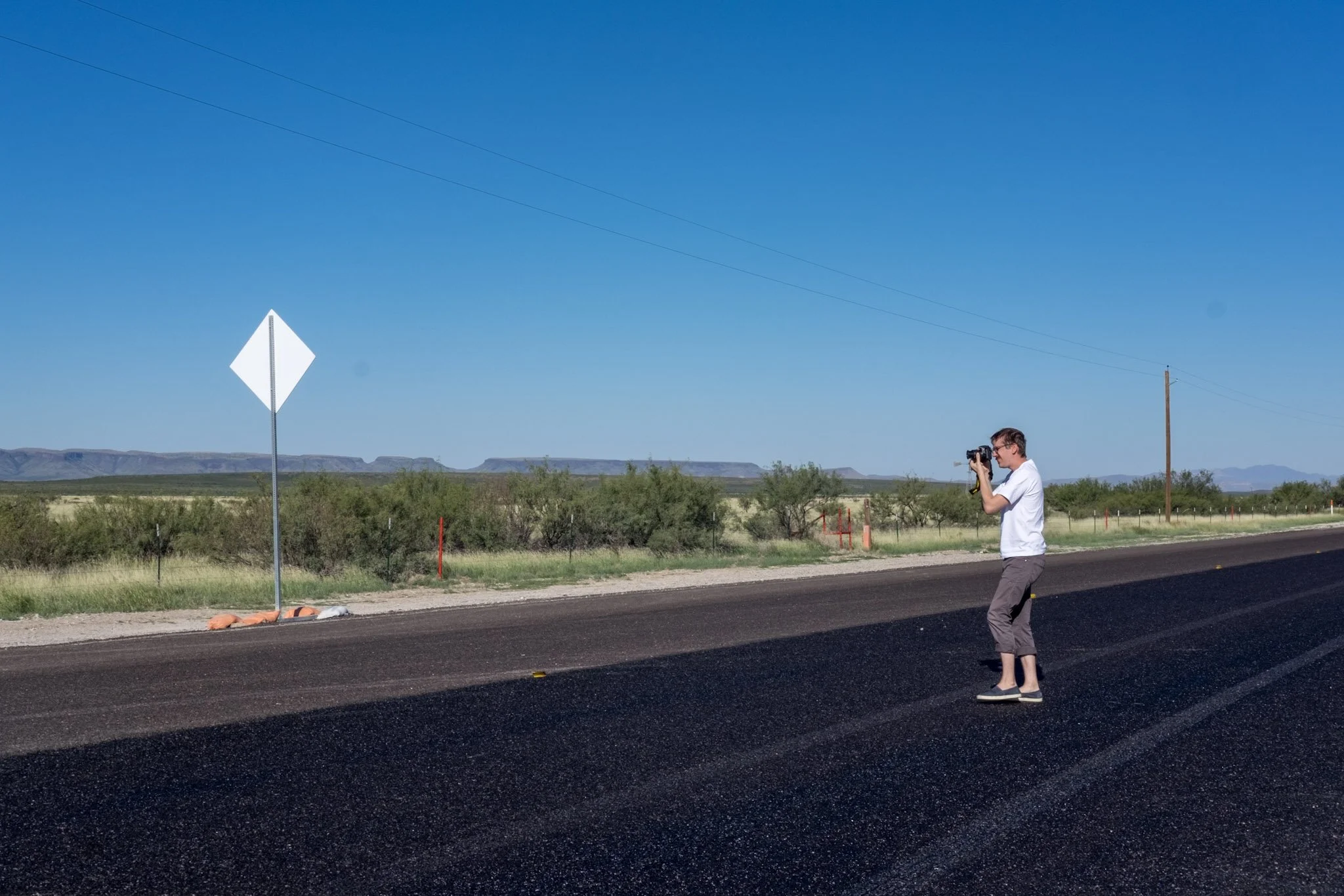 A man with glasses and a white shirt standing on the side of a road, looking through binoculars. The background shows a desert landscape with mountains and a clear blue sky. There are utility poles and sparse vegetation along the road.