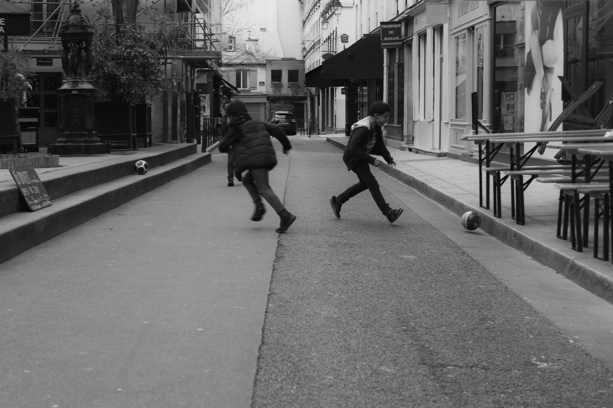 Two children playing soccer on a city street, with buildings and storefronts on either side, in black and white.