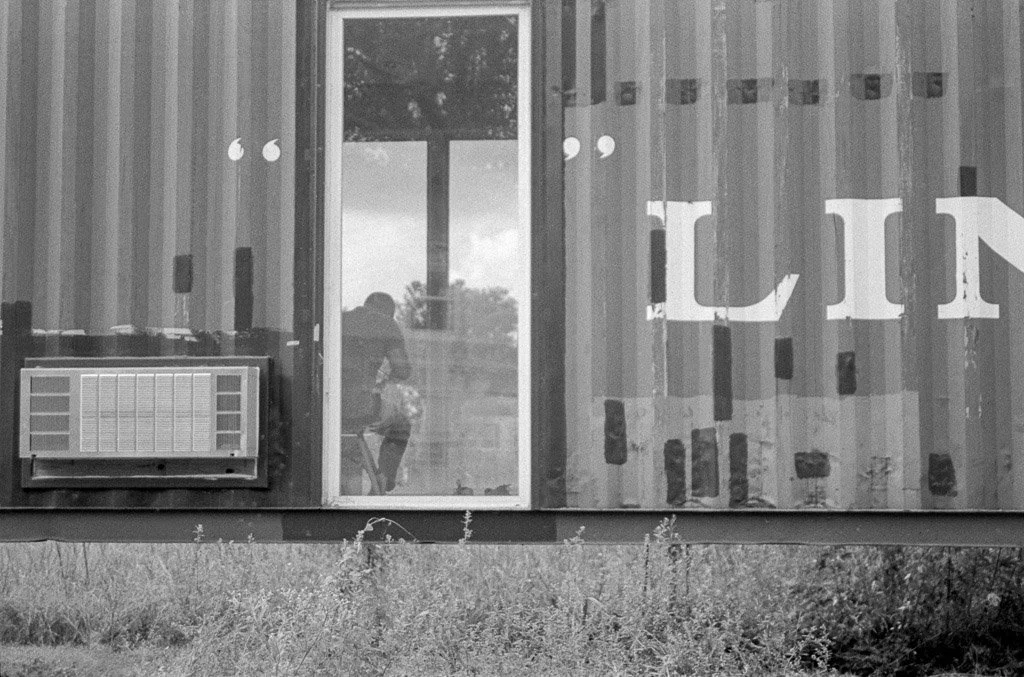Reflection of a person sitting and working inside a building with large windows, air conditioning unit, and partial text 'LIT' on the exterior wall.