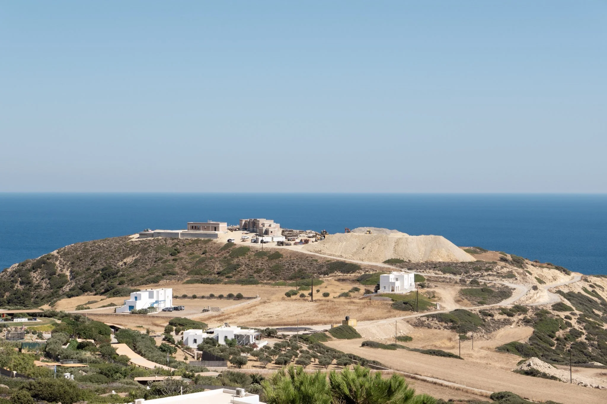 Landscape view of a coastal area with white houses, green trees, dry fields, and a hill leading to the blue sea and clear sky in the background.