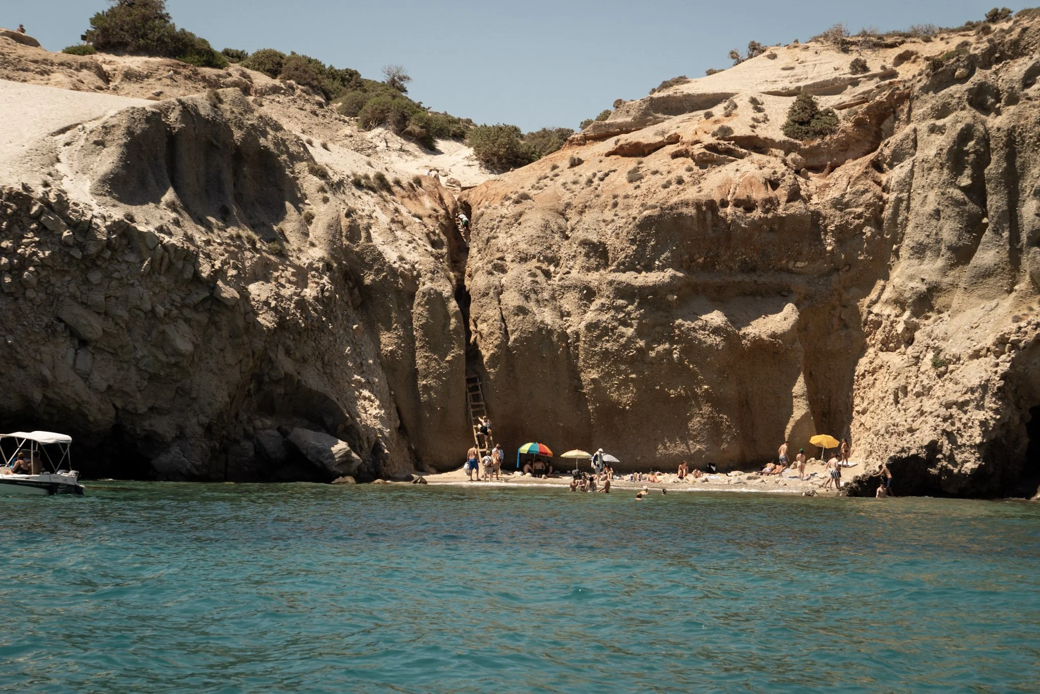 Beach scene with large rocky cliffs, people under umbrellas, and a boat on clear blue water.