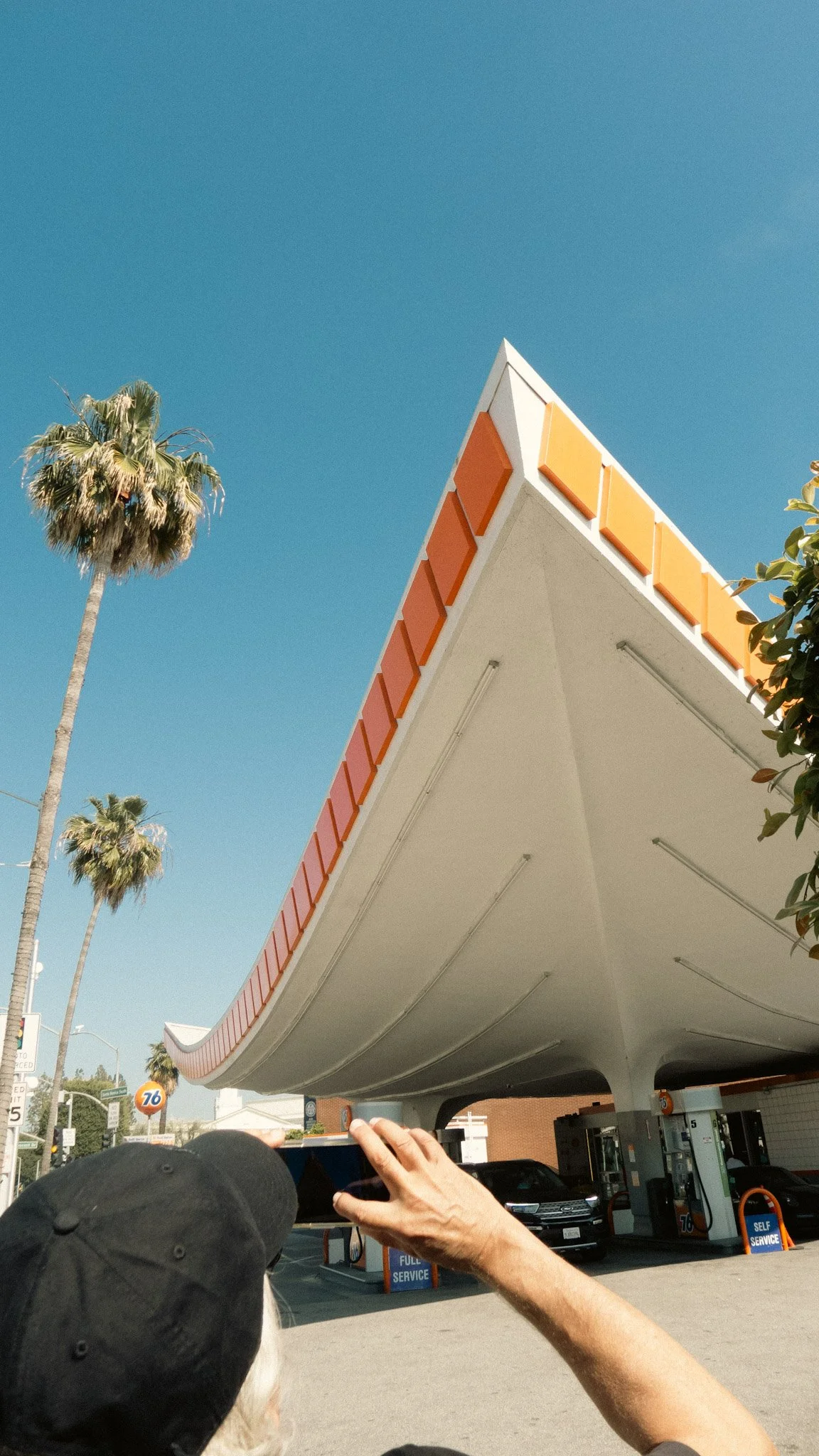 Person taking a photo of a building with a distinctive angled roof near a gas station with palm trees in the background.