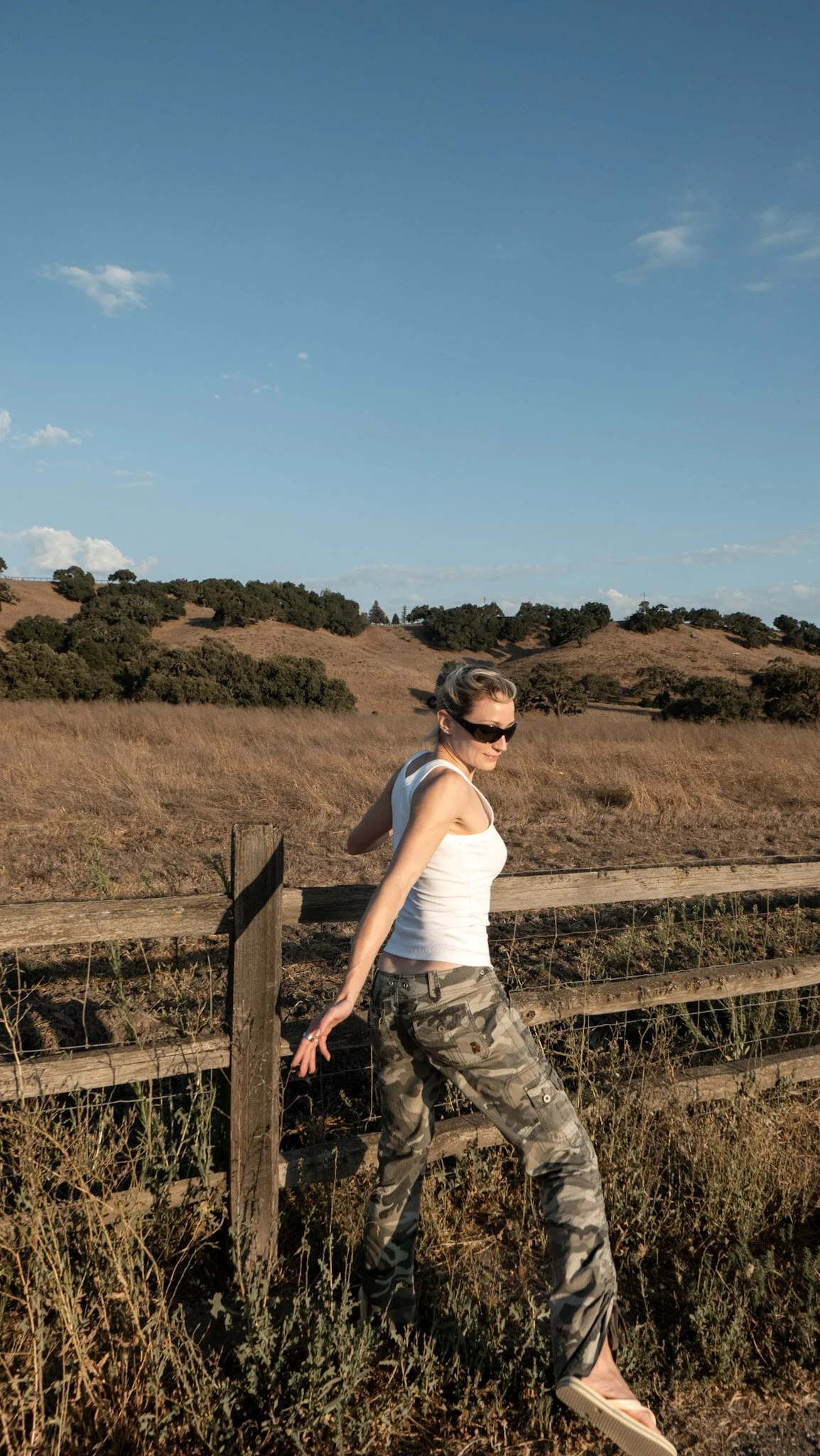 A woman wearing sunglasses, a white tank top, camouflage pants, and sandals standing outdoors near a wooden fence, with a landscape of rolling hills and a partly cloudy sky in the background.