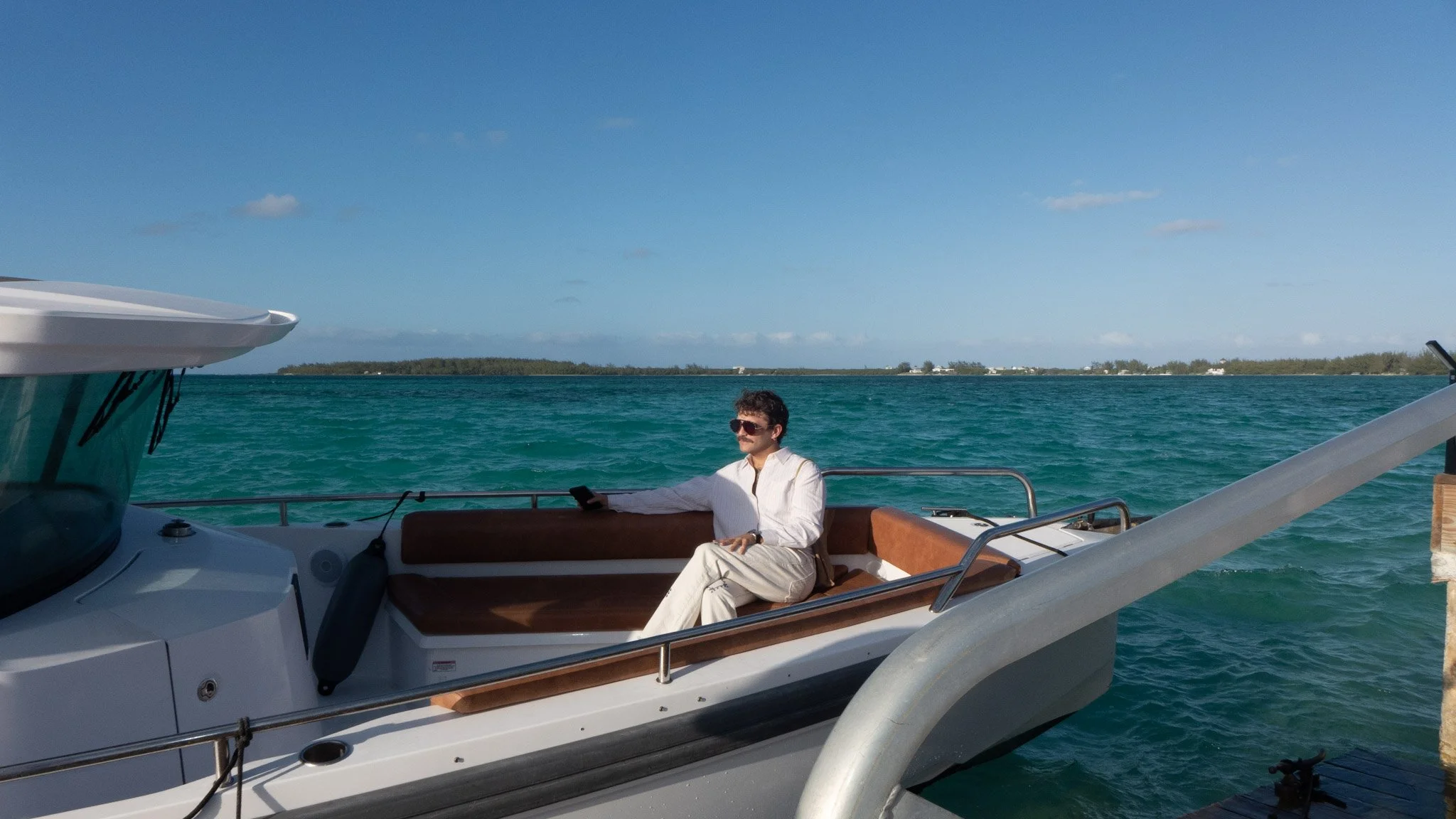 Man in sunglasses and white shirt sitting on a lounge chair on a yacht, holding a smartphone, with blue ocean water and small islands in the background under a partly cloudy sky.