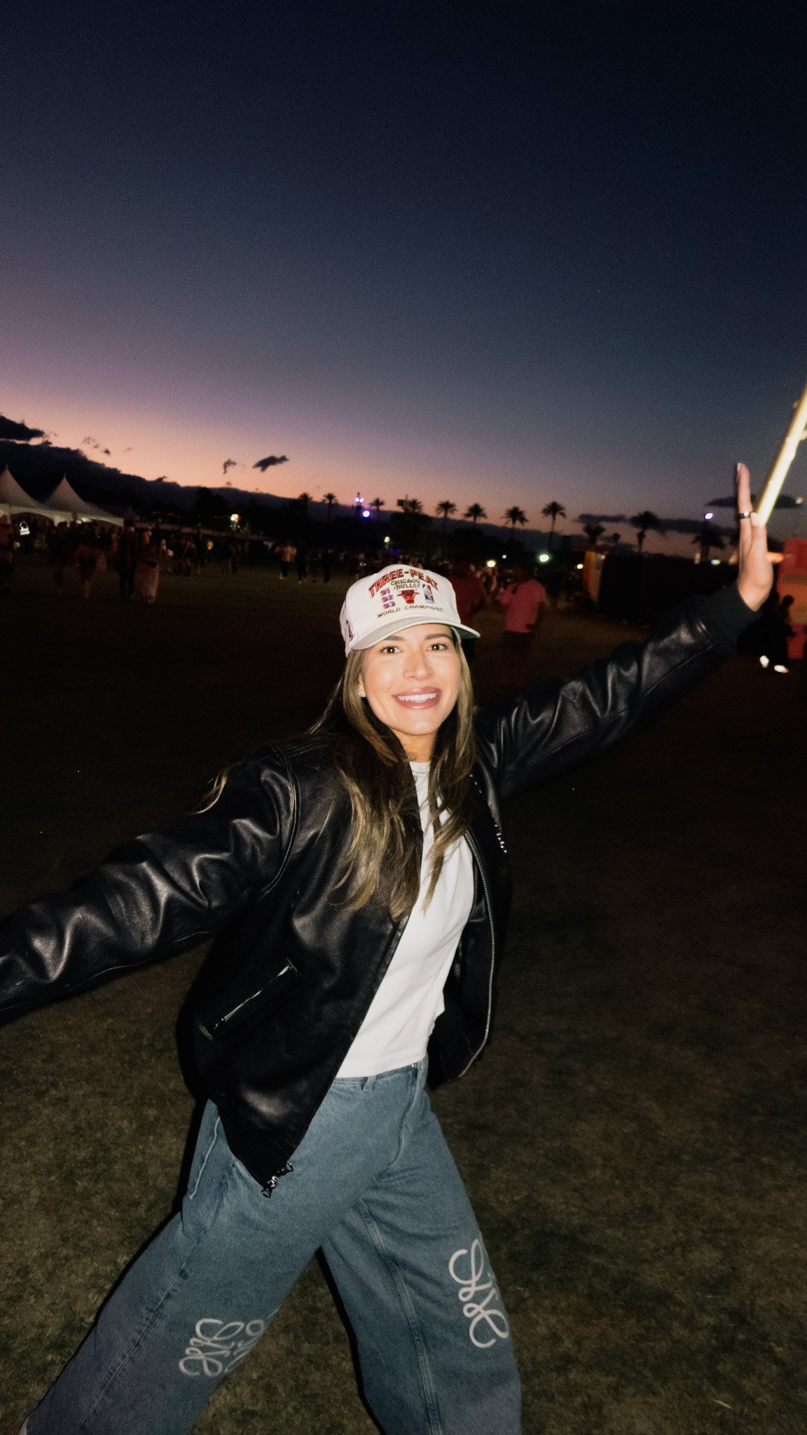 A woman smiling and dancing outdoors at dusk, wearing a white hat, black leather jacket, white shirt, and grey jeans, with a festive event and tent in the background.