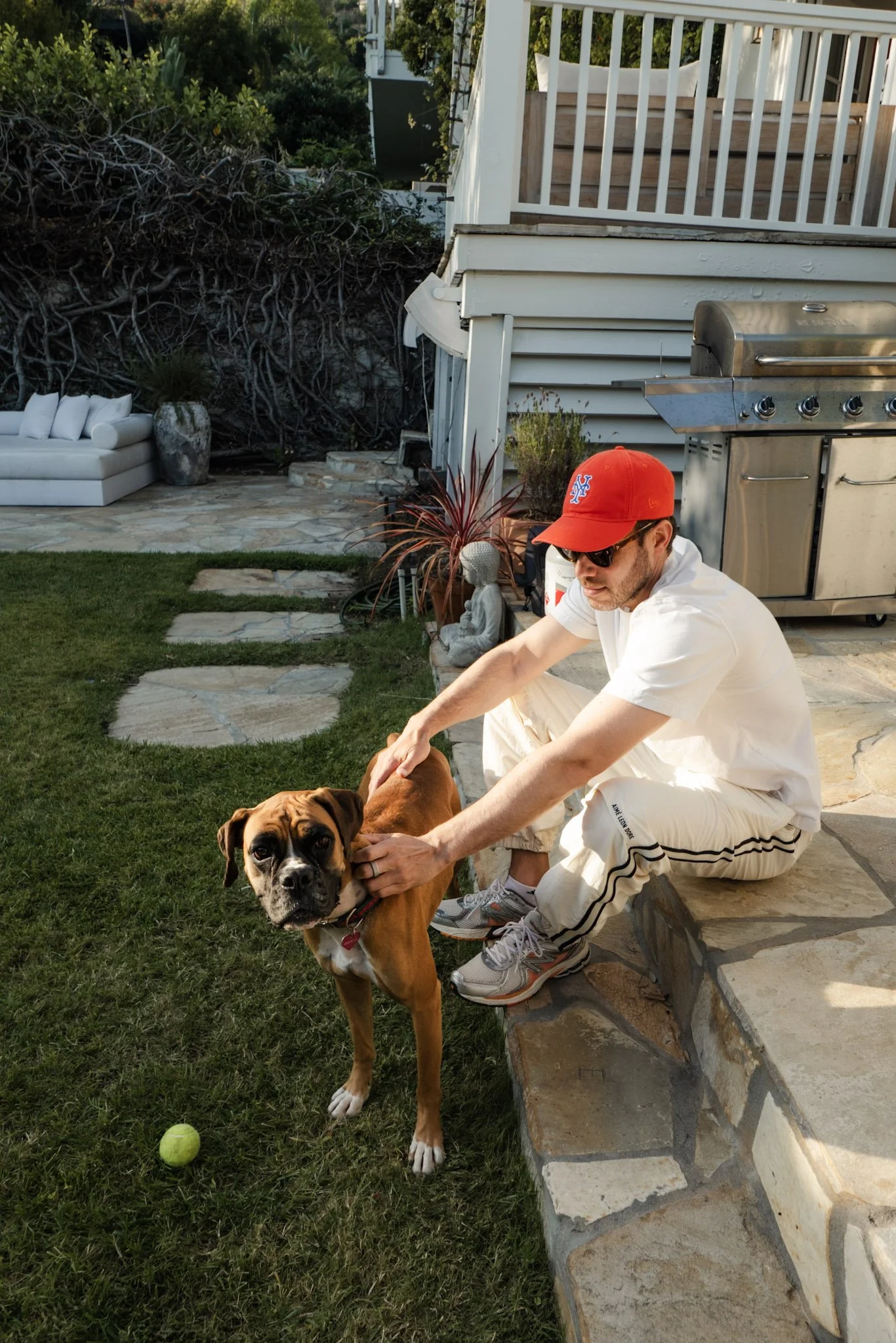 A man in a white shirt, white pants, sunglasses, and a red New York Mets cap is kneeling on a stone patio, holding a brown and white dog with a black face. The dog is standing on the grass near a tennis ball. The background shows a deck with white ra