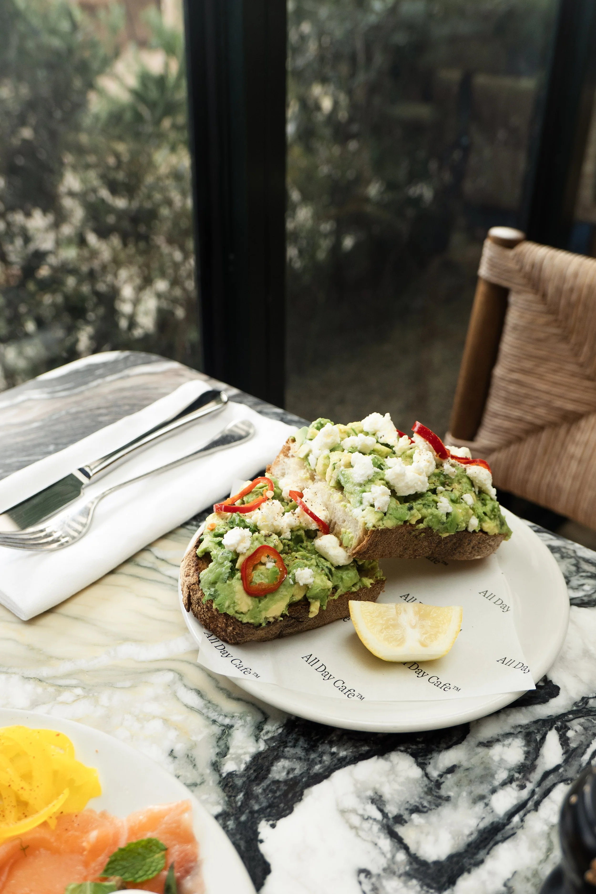 A plate of avocado toast topped with crumbled cheese and sliced red chili peppers, served with a lemon wedge, on a marble table at a cafe with a view of trees outside.