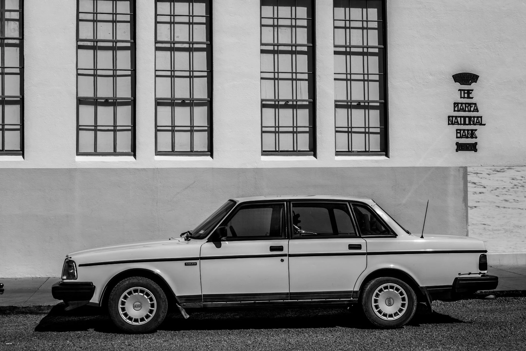 A vintage white Volvo sedan parked in front of a white building with multiple large windows and a sign that reads 'The Marfa National Bank'.