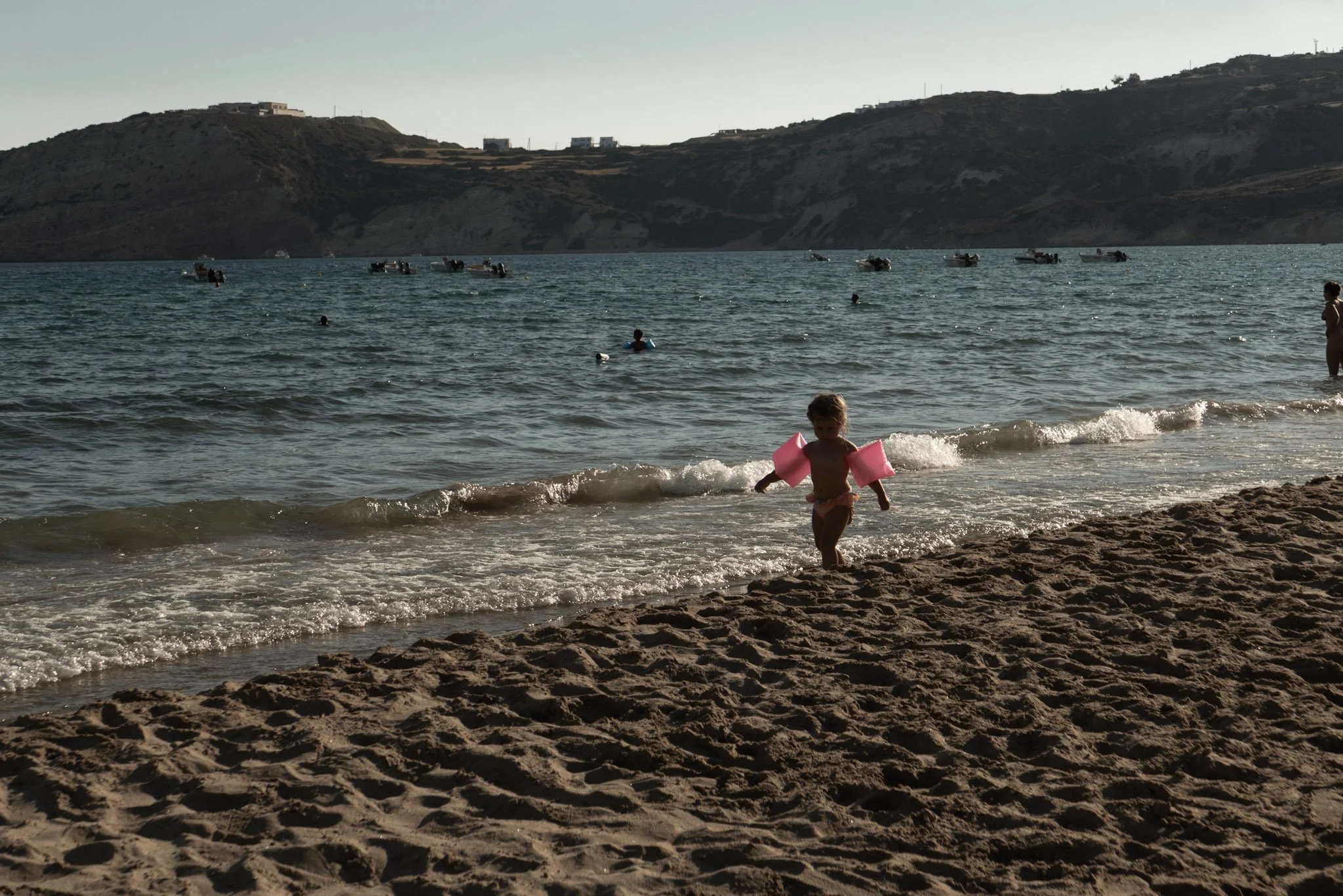 A young child with pink floaties walking on the sandy beach near the shoreline, with people swimming in the water and boats floating on the surface, and hills in the background.