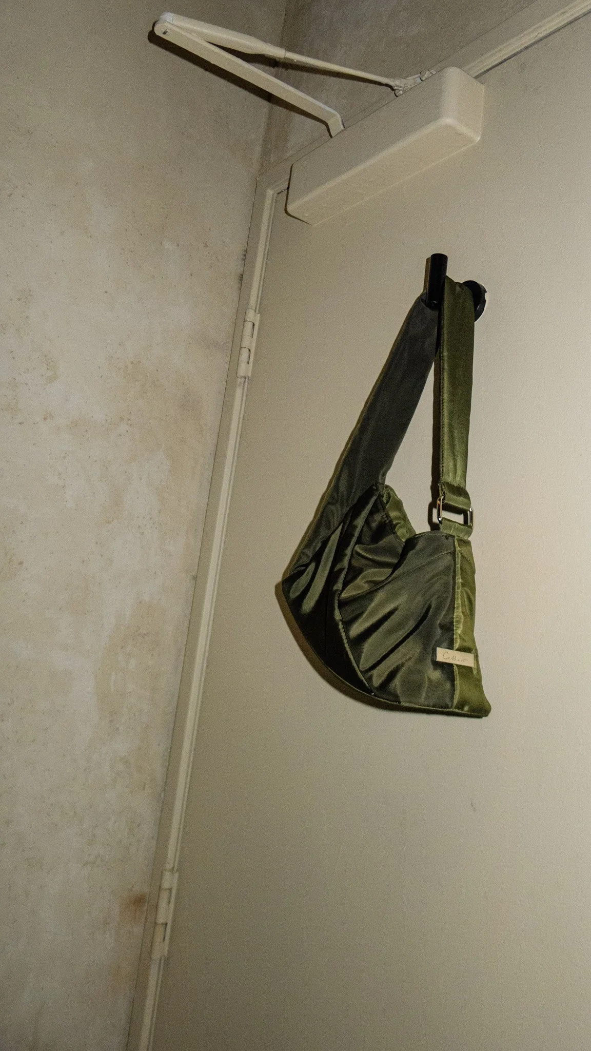 A green camouflage-style handbag hanging from a black hook on a white wall, near a beige door with a white door frame.