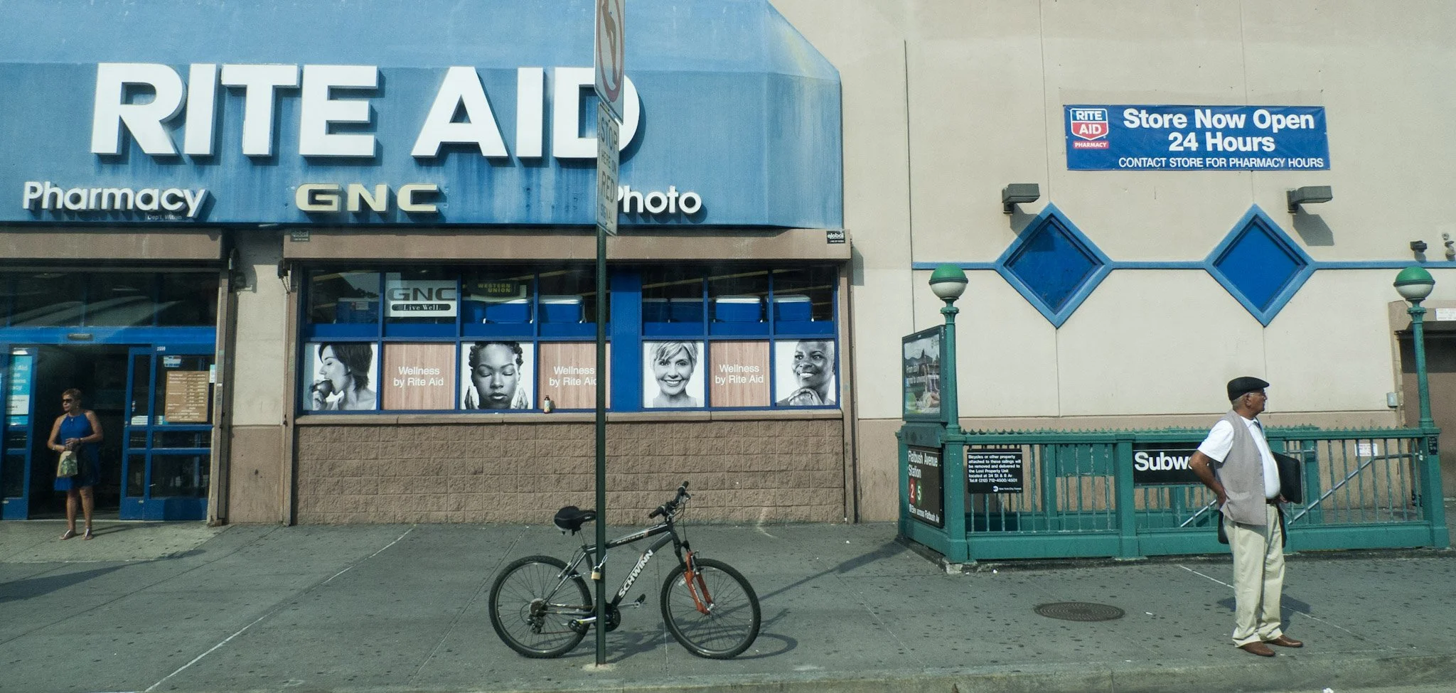 The exterior of a Rite Aid pharmacy store with a large blue sign that reads 'Store Now Open 24 Hours' and a smaller purple sign indicating pharmacy hours. There are two pedestrians visible: a woman in a blue dress near the entrance, and a man in a wh