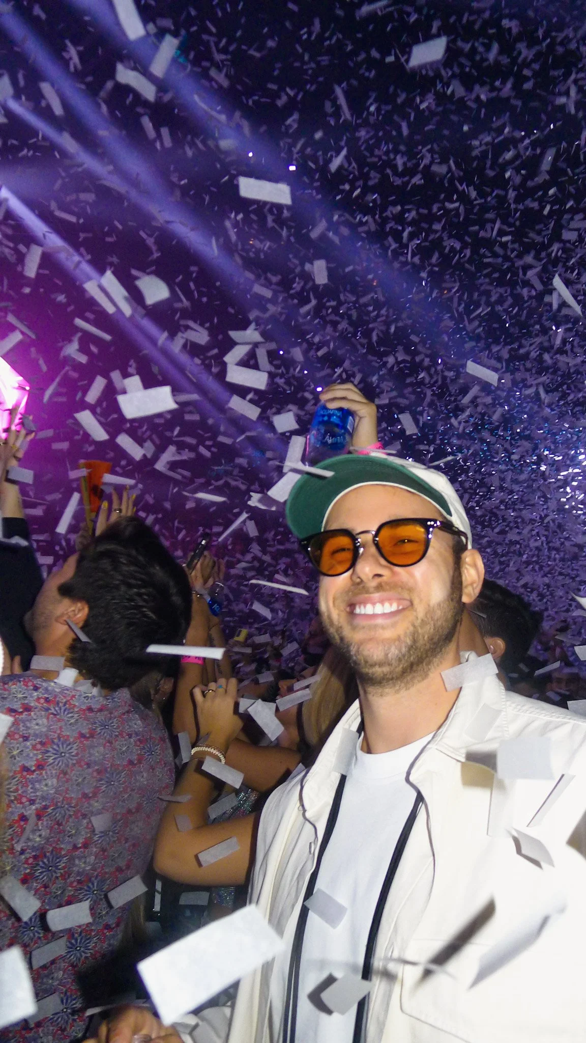 A man smiling at a party, wearing sunglasses and a white jacket with black trim, surrounded by others, confetti falling from above, and purple lighting.