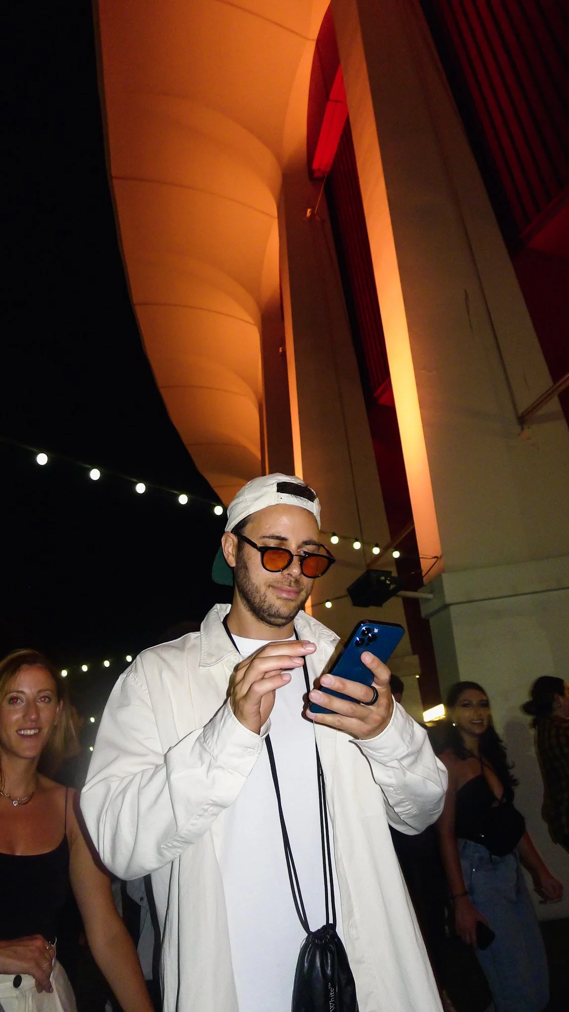 A man wearing a white jacket, sunglasses, and a backwards cap, is looking at his phone while standing at a social event with people and string lights in the background.