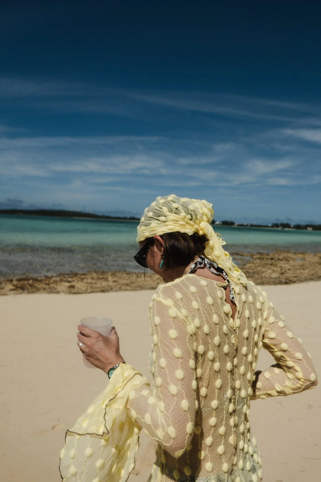 A woman in a yellow dress with pom-pom details walking on a sandy beach, holding a cup, with water and sky in the background.