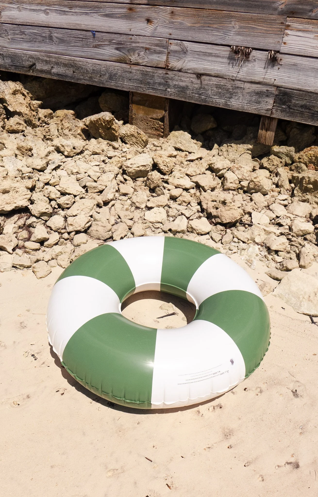 A green and white striped inflatable pool float on sandy beach with rocks and wooden structure in background.