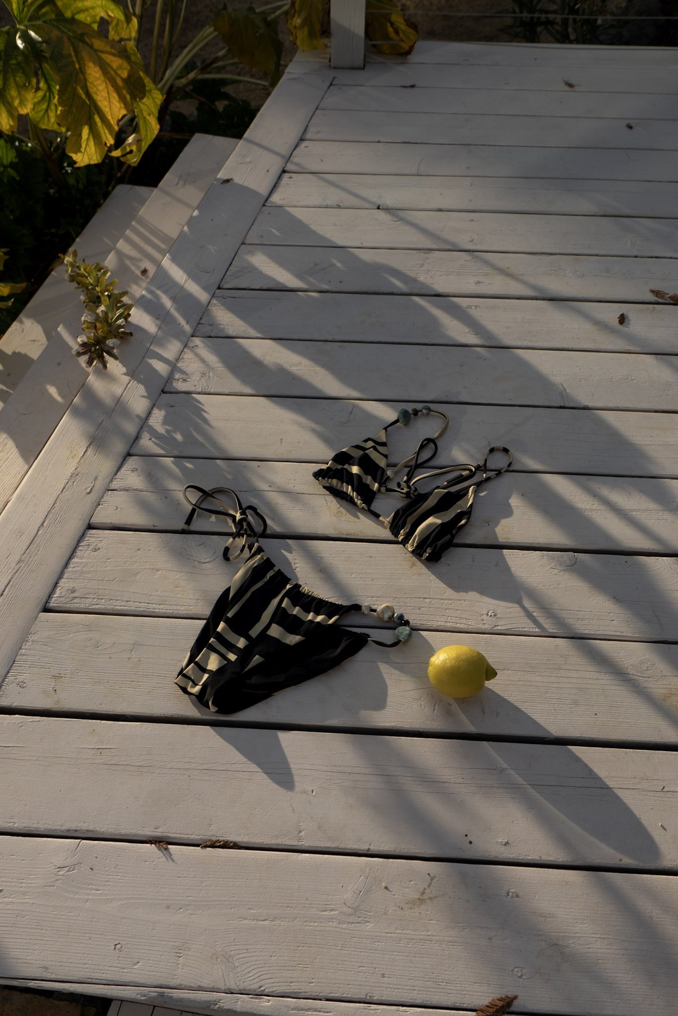 A white wooden outdoor deck with a yellow lemon, black and white striped bikini, a beaded necklace, and a leaf attached to a plant in the corner. Shadows from a railing and nearby foliage are cast on the deck.