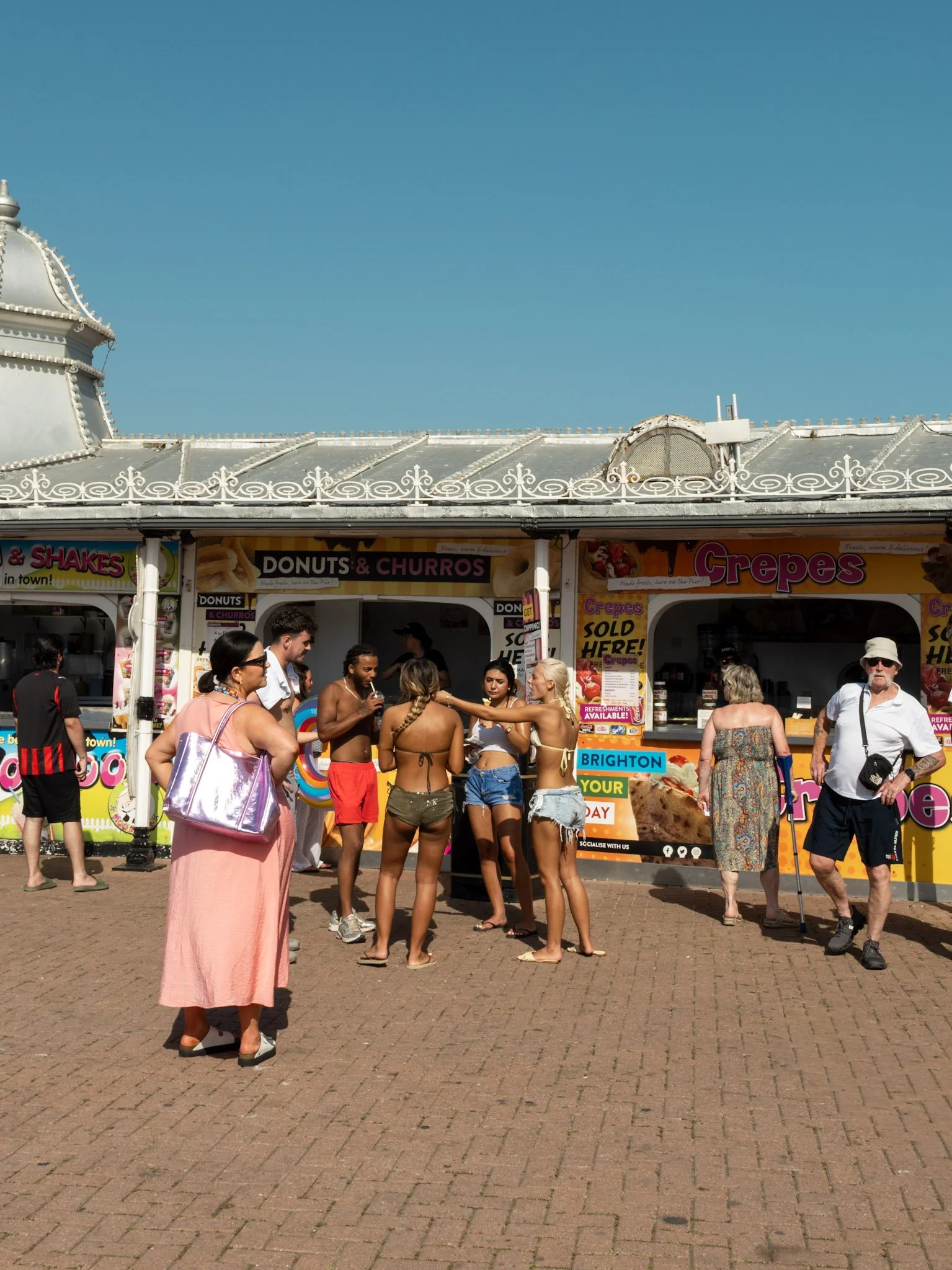 A group of people gathered around a food stand at the Brighton seaside, with some people wearing swimsuits and summer clothes, under a clear blue sky.
