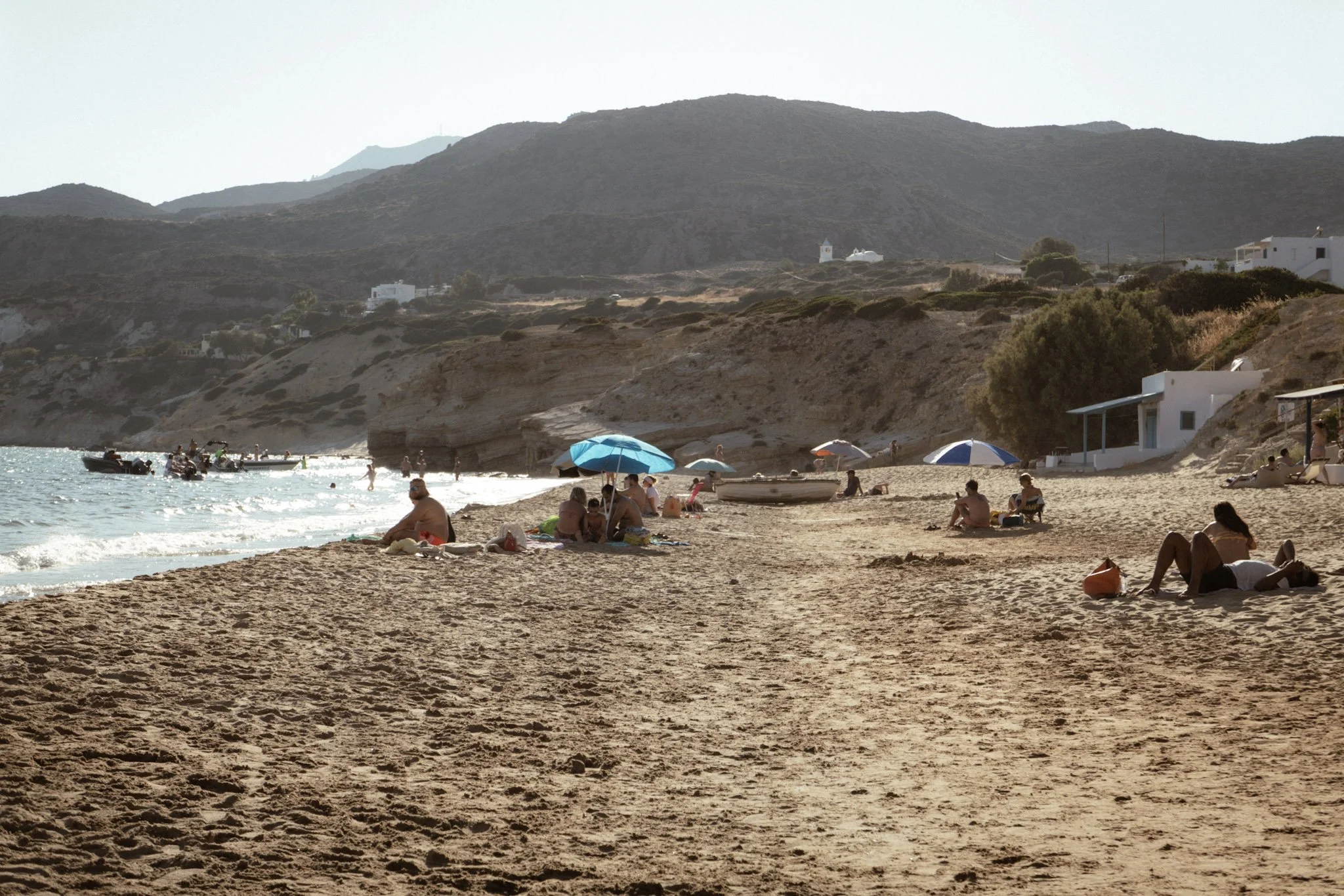 People relaxing on a sandy beach with umbrellas and boats, mountainous landscape in the background.