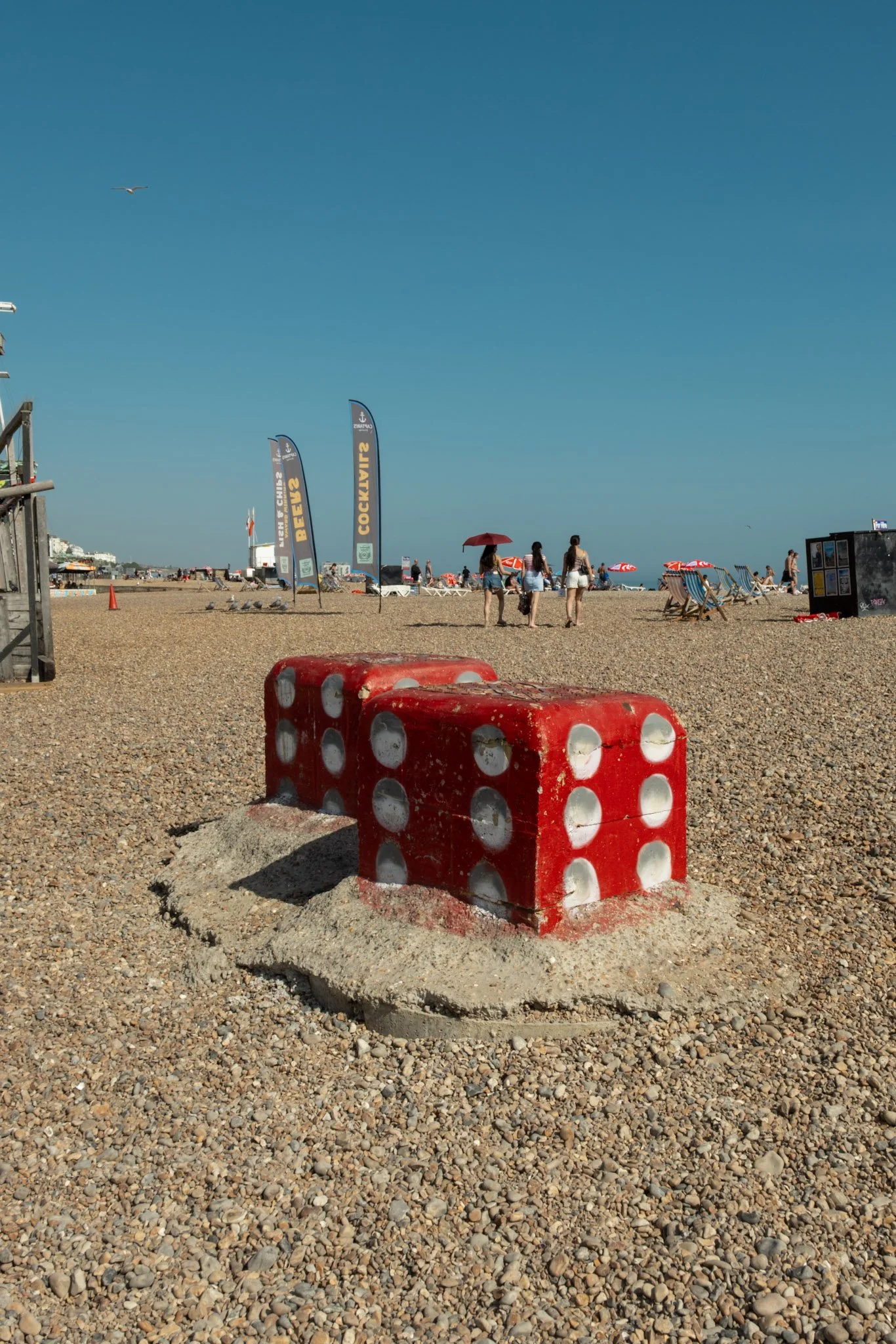 A large red and white dice-shaped barrier on a pebble beach with people, umbrellas, flags, and the ocean in the background on a sunny day.