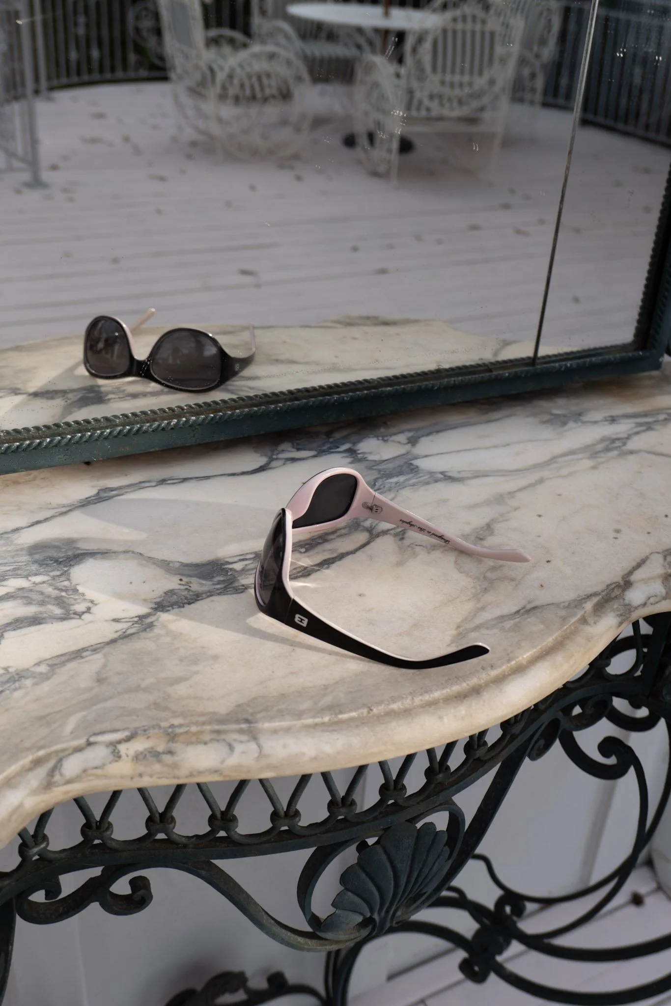 A pair of sunglasses on a marble table with a window reflecting an outdoor patio with white furniture and a railing.