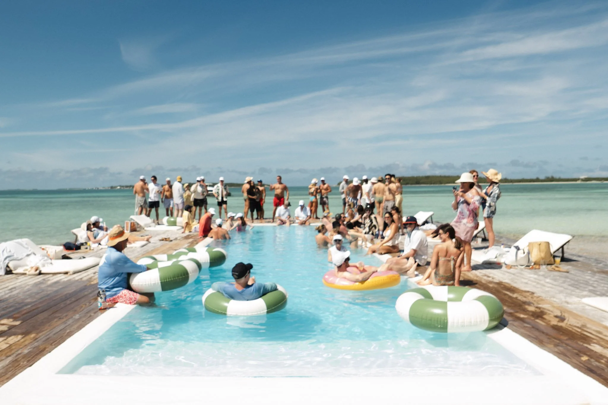 People gathered around a swimming pool on a wooden deck, some sitting and some standing, with beach balls and pool floats, under a blue sky with some clouds.
