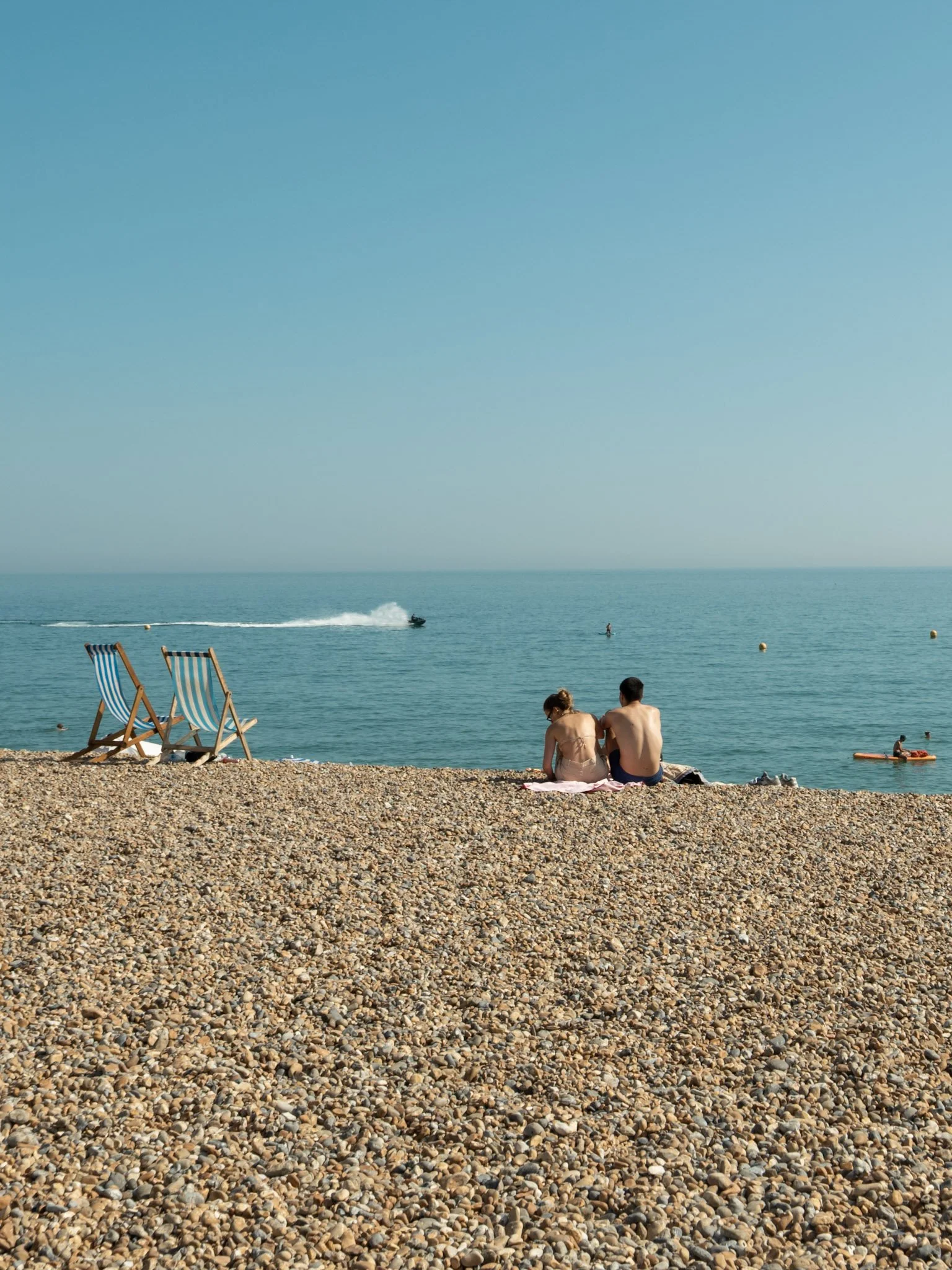 A pebble beach with two striped deck chairs, a couple sitting on the shore, and people swimming and kayaking in the water. The sky is clear and blue.