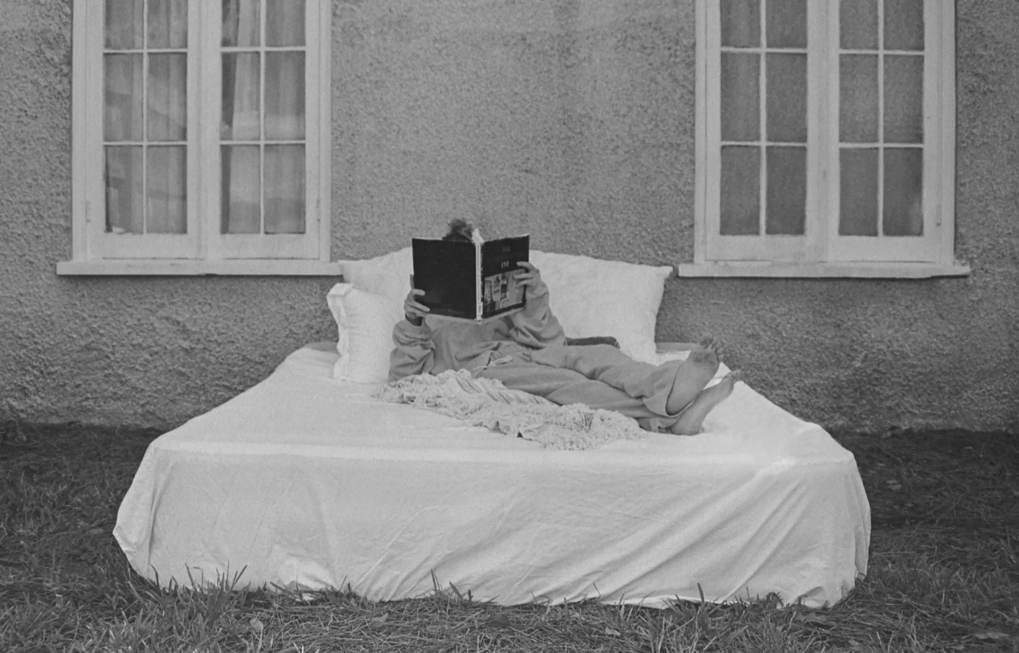 A person lies on a bed reading a book outside a house with two windows, in black-and-white.