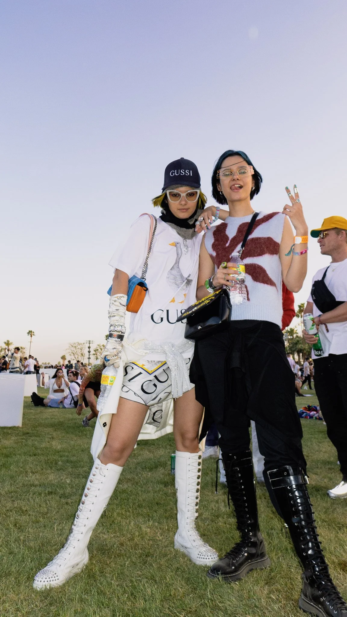 Two fashionable young women posing at an outdoor event, standing close together. One wears a white ensemble with knee-high lace-up boots, sunglasses, and a black cap that says "GUSSI". The other wears a sleeveless sweater with bold red and white patt