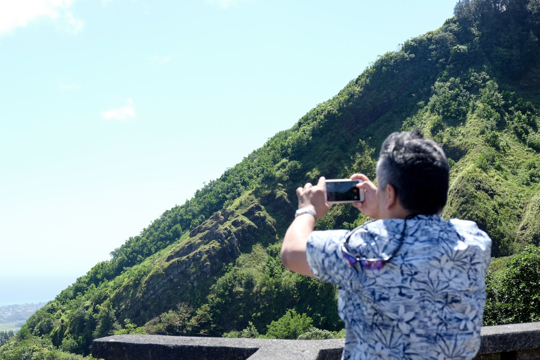 A man taking a photo with a smartphone of a lush green hillside with trees and a blue sky in the background.
