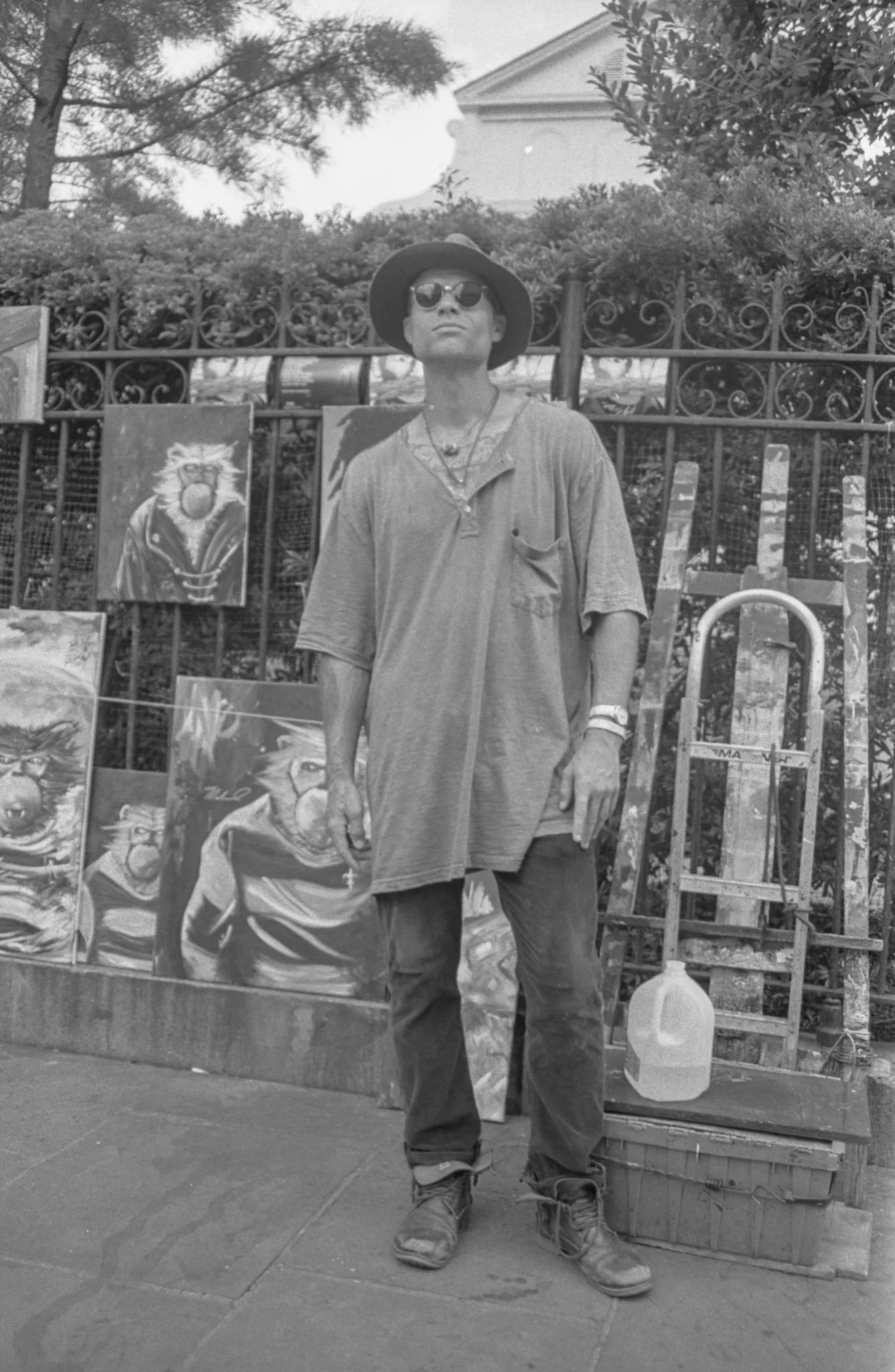 A man wearing a wide-brimmed hat, sunglasses, and casual clothing, standing on a sidewalk next to an artist's display of paintings of lions. A large plastic container with a drink is at his feet. Behind him is a fence with trees and a house in the ba