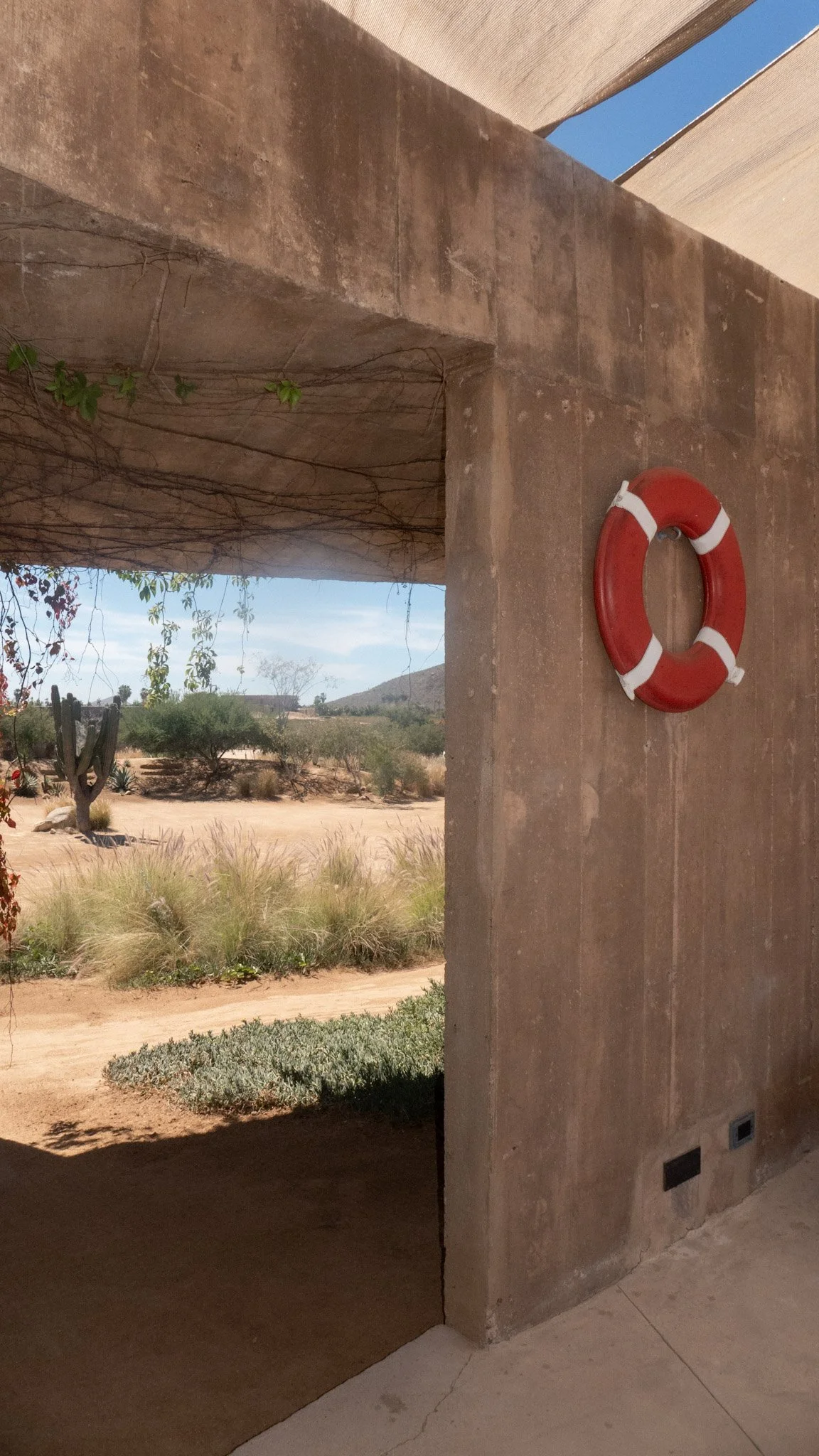 A red and white lifebuoy hangs on a concrete wall underneath a shaded area, with a desert landscape featuring bushes, cacti, and distant mountains visible beyond.