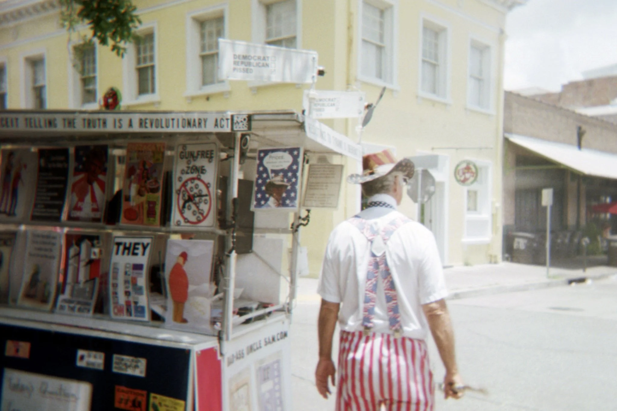 A street scene with a man walking past a newsstand covered with various posters and signs, including one that reads 'GUN FREE ZONE' and another indicating political messages. The man is dressed in a white shirt with a patterned vest and red-striped s