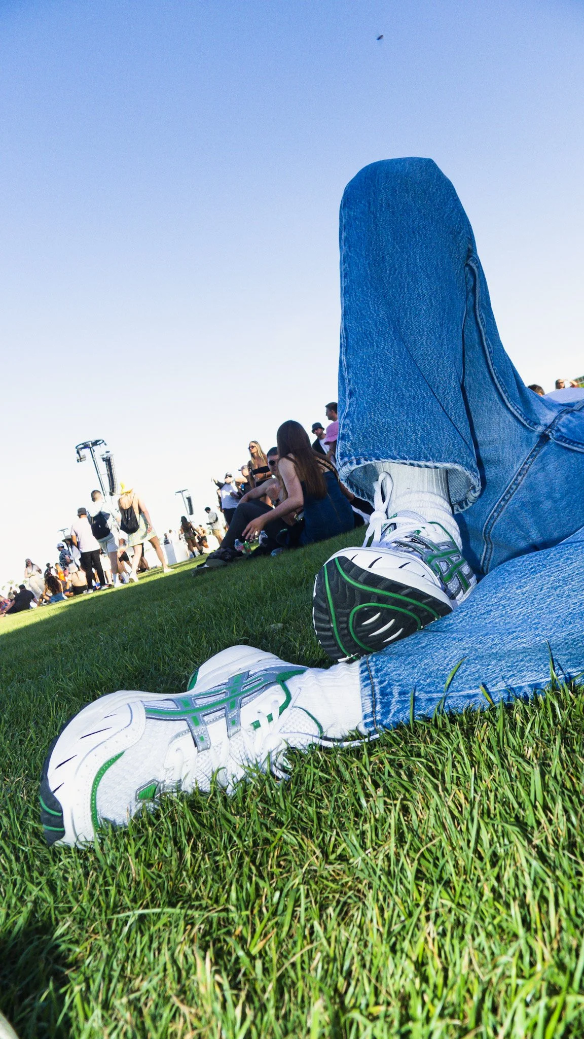 View of a person lying on grass at an outdoor event, with their legs stretched out and wearing white sneakers and blue jeans. Around them are other people sitting and standing, with a clear blue sky overhead.