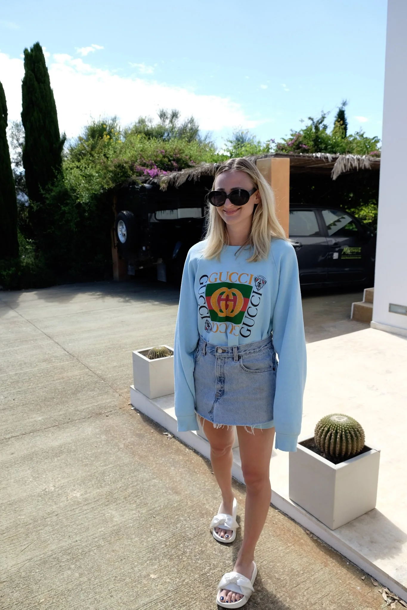 A young woman with blonde hair wearing sunglasses, a light blue Gucci sweatshirt with a colorful logo, a denim skirt, and white slippers with fur trim, standing outdoors on a sunny day with a clear blue sky, palm trees, and greenery in the background