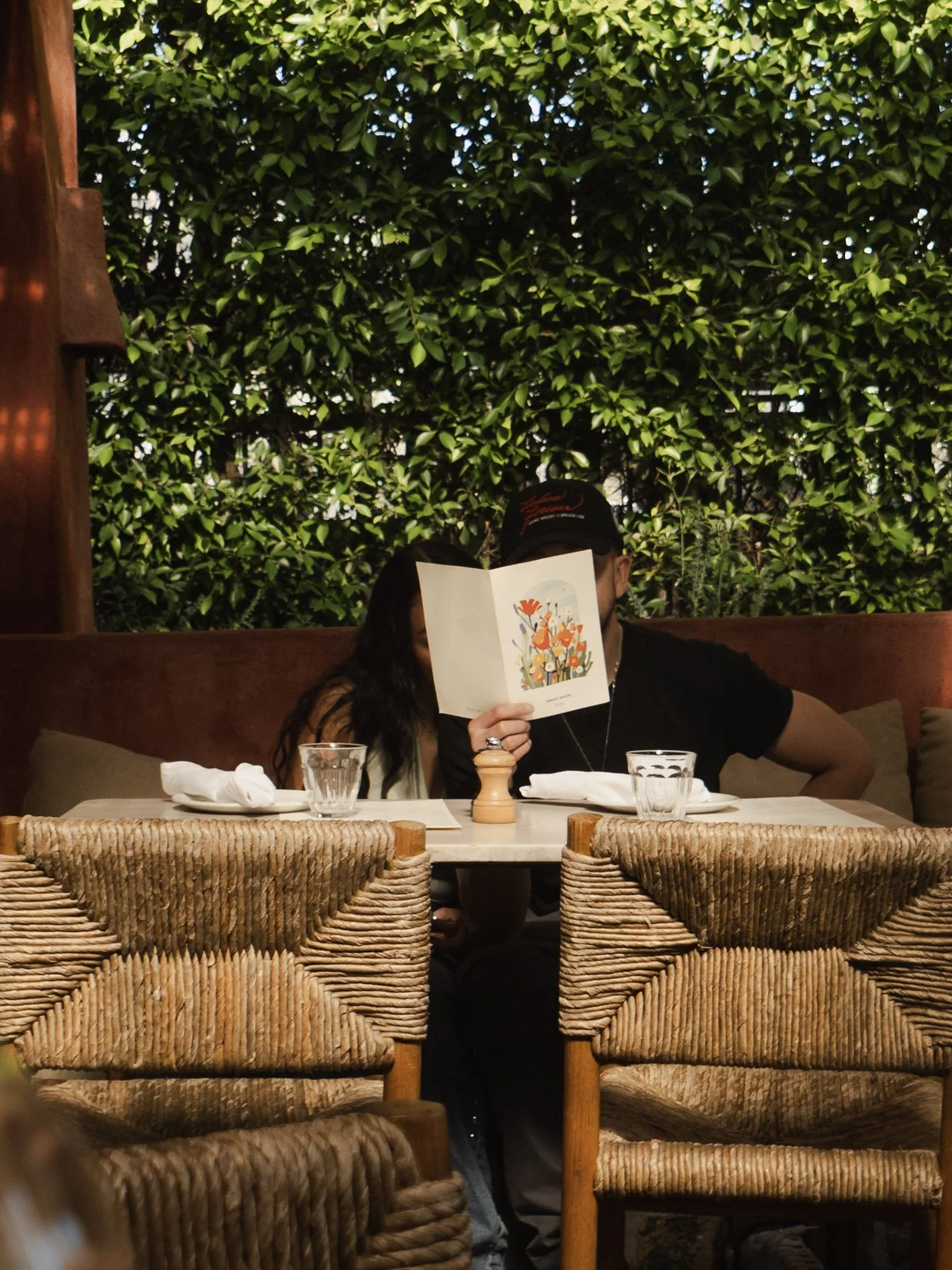 People sitting at a restaurant table with wicker chairs, one person reading a menu, surrounded by greenery in a cozy dining setting.