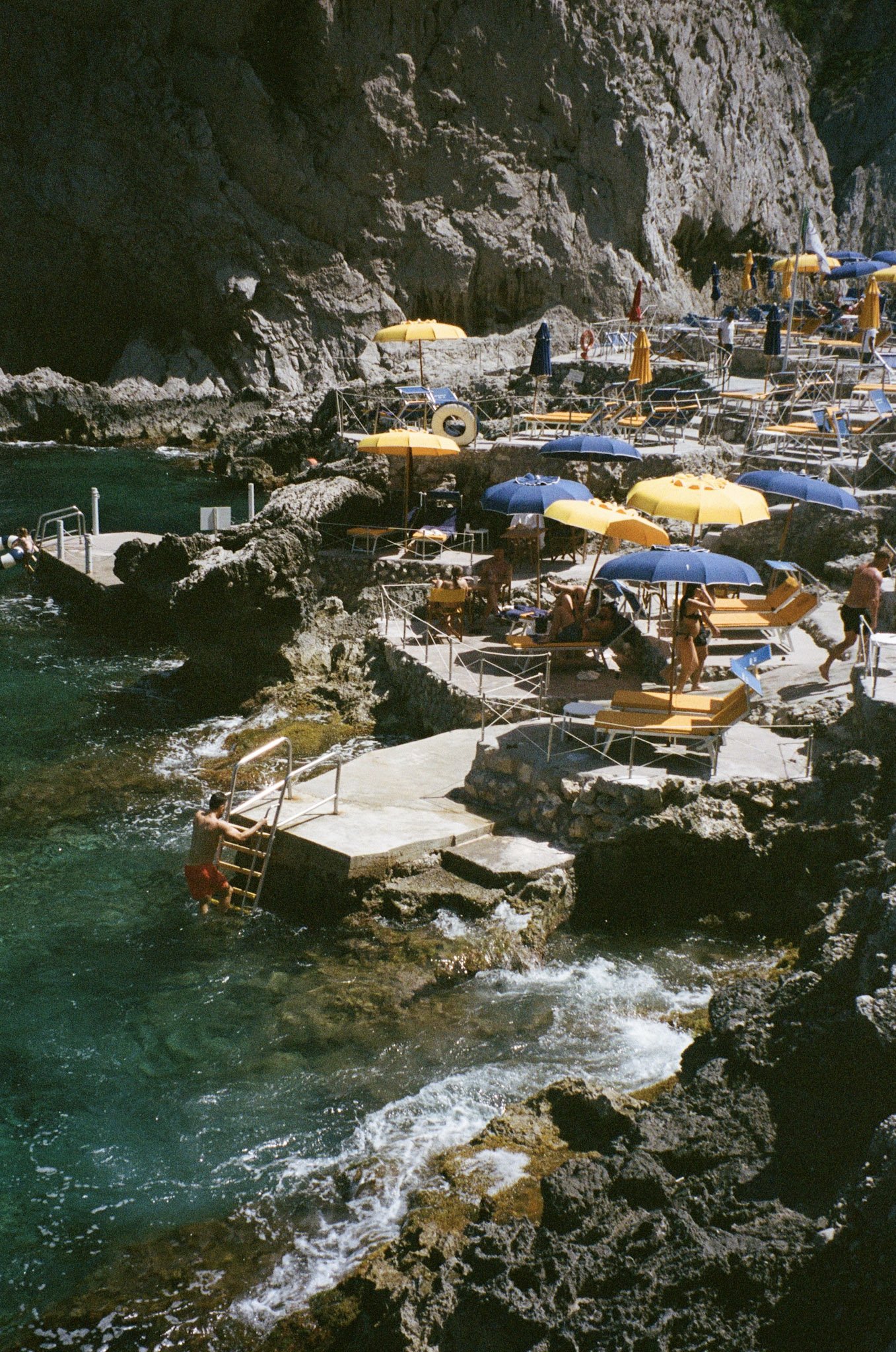 People swimming and relaxing on rocky platforms near a rocky cliffside with yellow and blue umbrellas and chairs in a coastal setting.