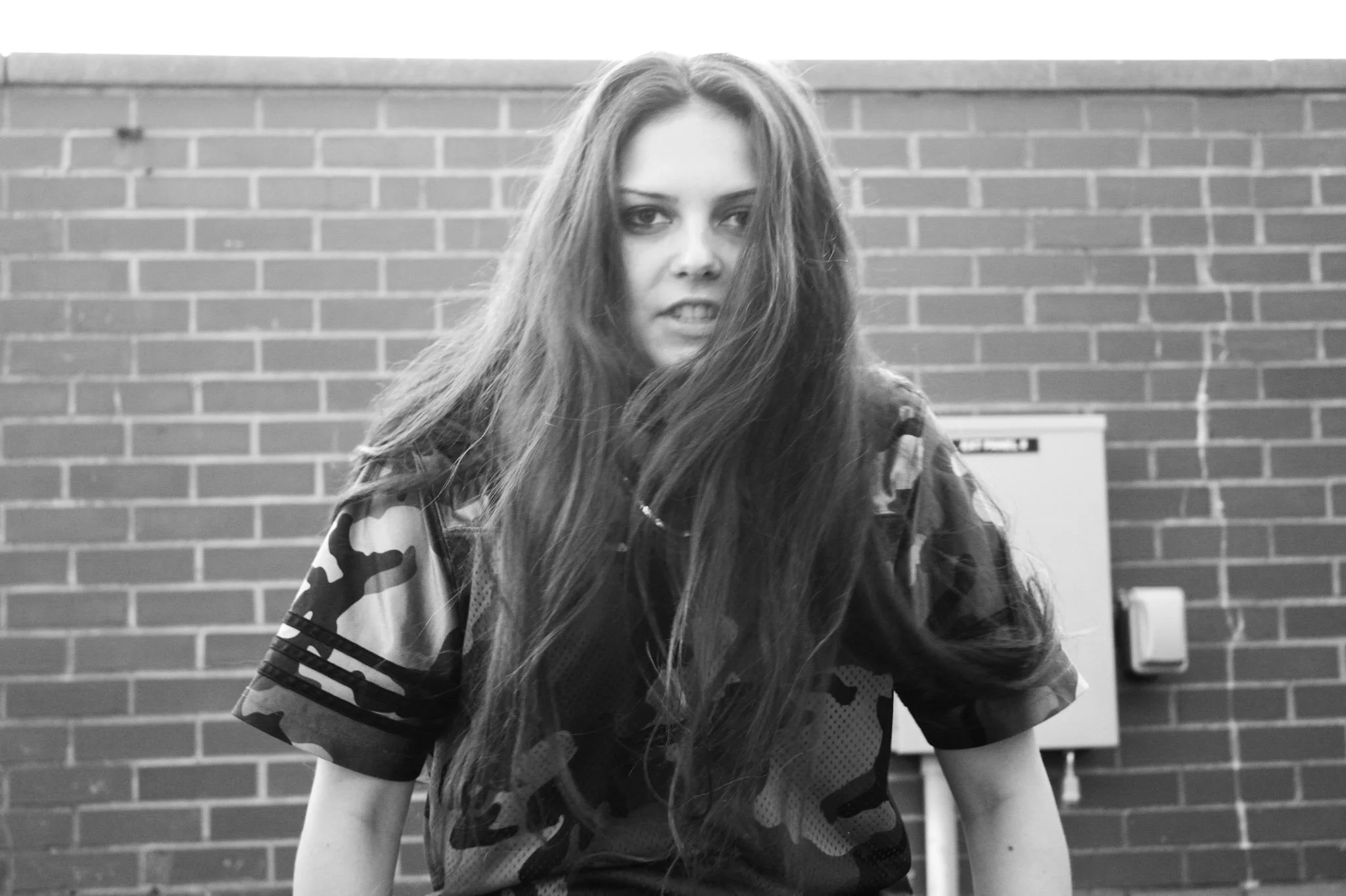 A young woman with long hair standing in front of a brick wall, wearing a sports jersey.
