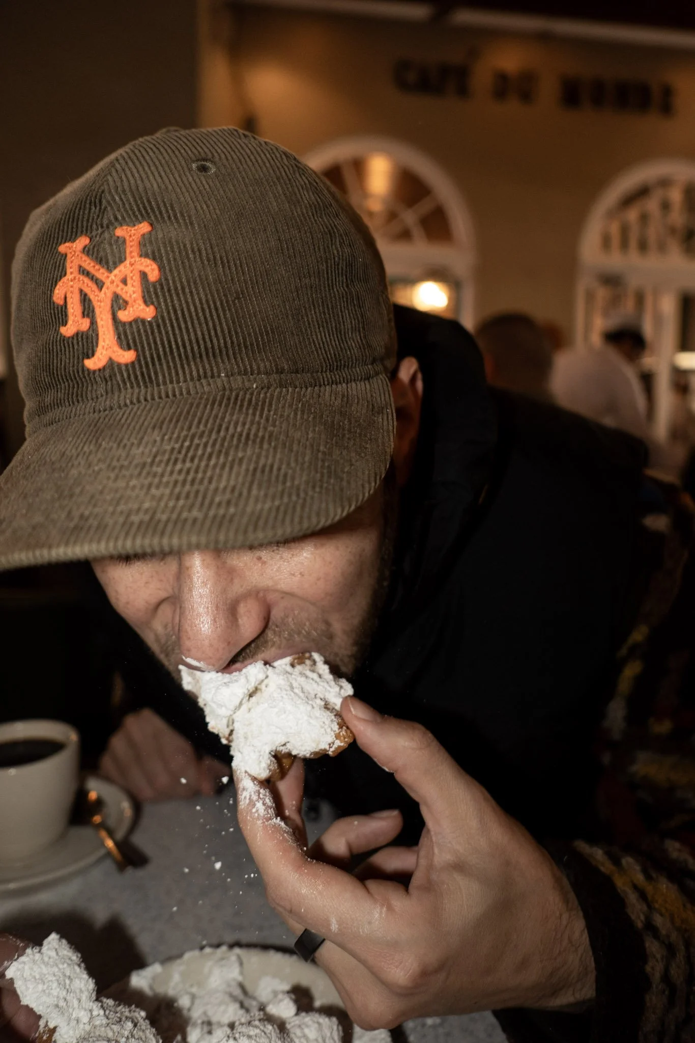 Person wearing a brown baseball cap with an orange New York Mets logo, holding a cake with white frosting, appearing to take a bite in a casual indoor setting with other people in the background.