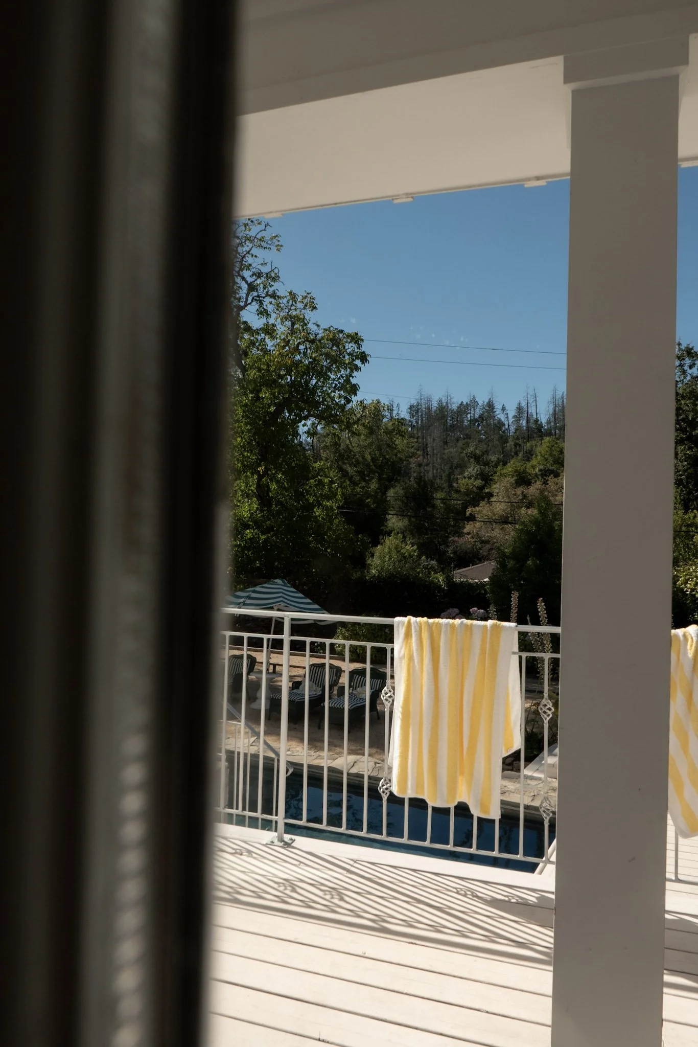 View of a backyard pool area seen through a window with vertical blinds, with yellow and white striped towel hanging on the railing, surrounded by green trees and a blue sky.