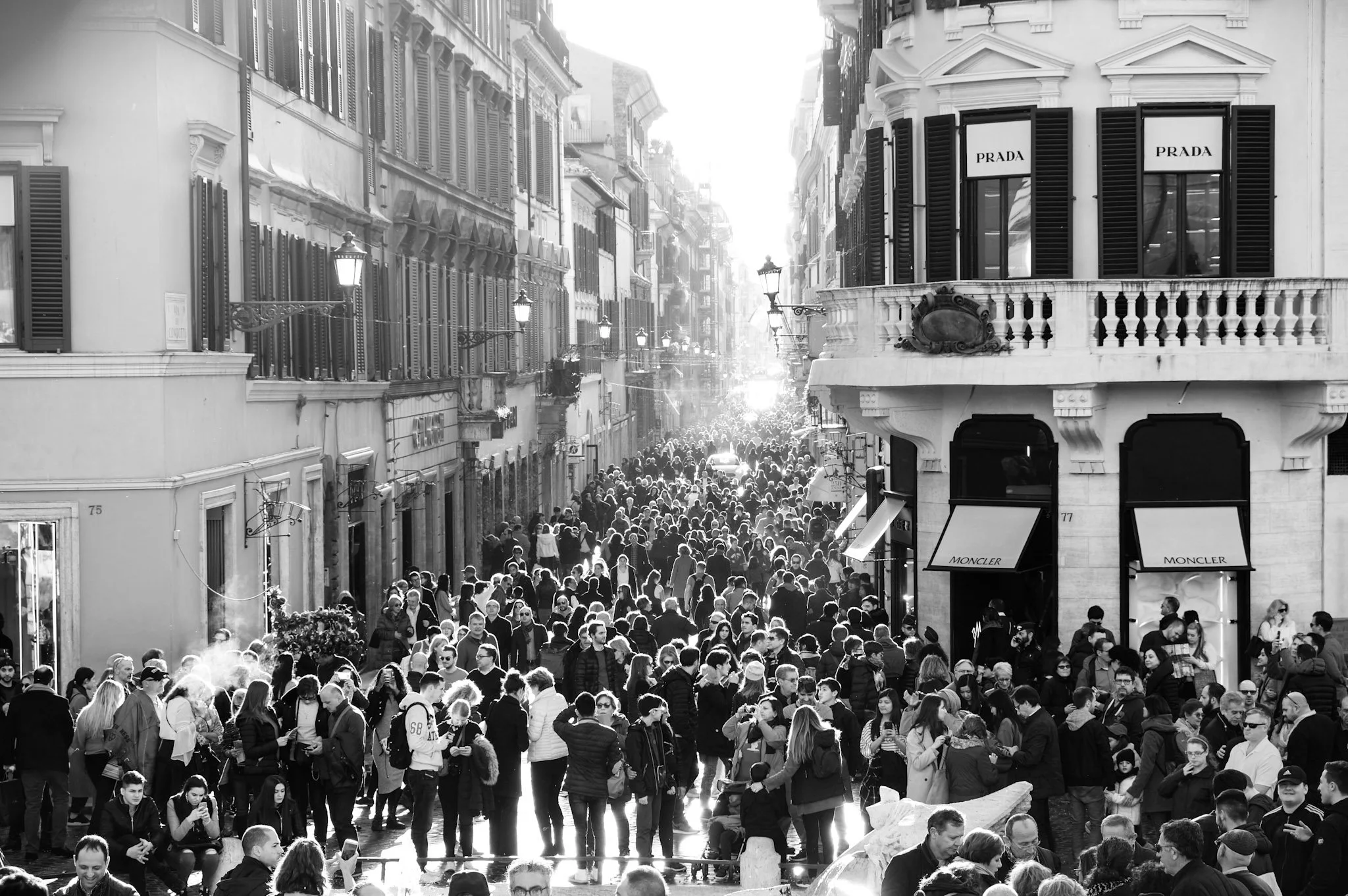 Black and white photo of a crowded street filled with people walking, with buildings on each side and signs from luxury brands like Prada and Moncler.