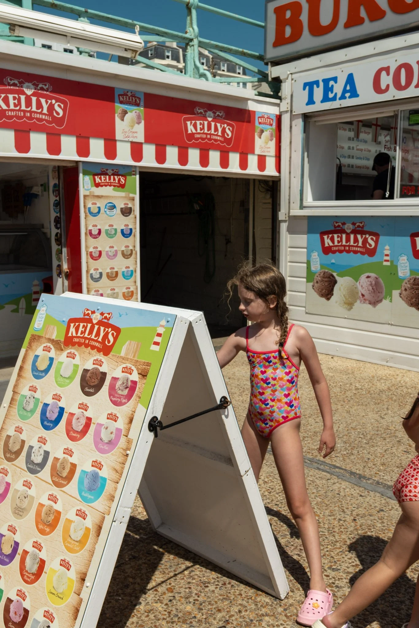 A food stand with signs advertising ice cream, tea, and cola, and a menu with various ice cream flavors. A girl in a colorful heart-patterned swimsuit and pink Crocs is walking by.