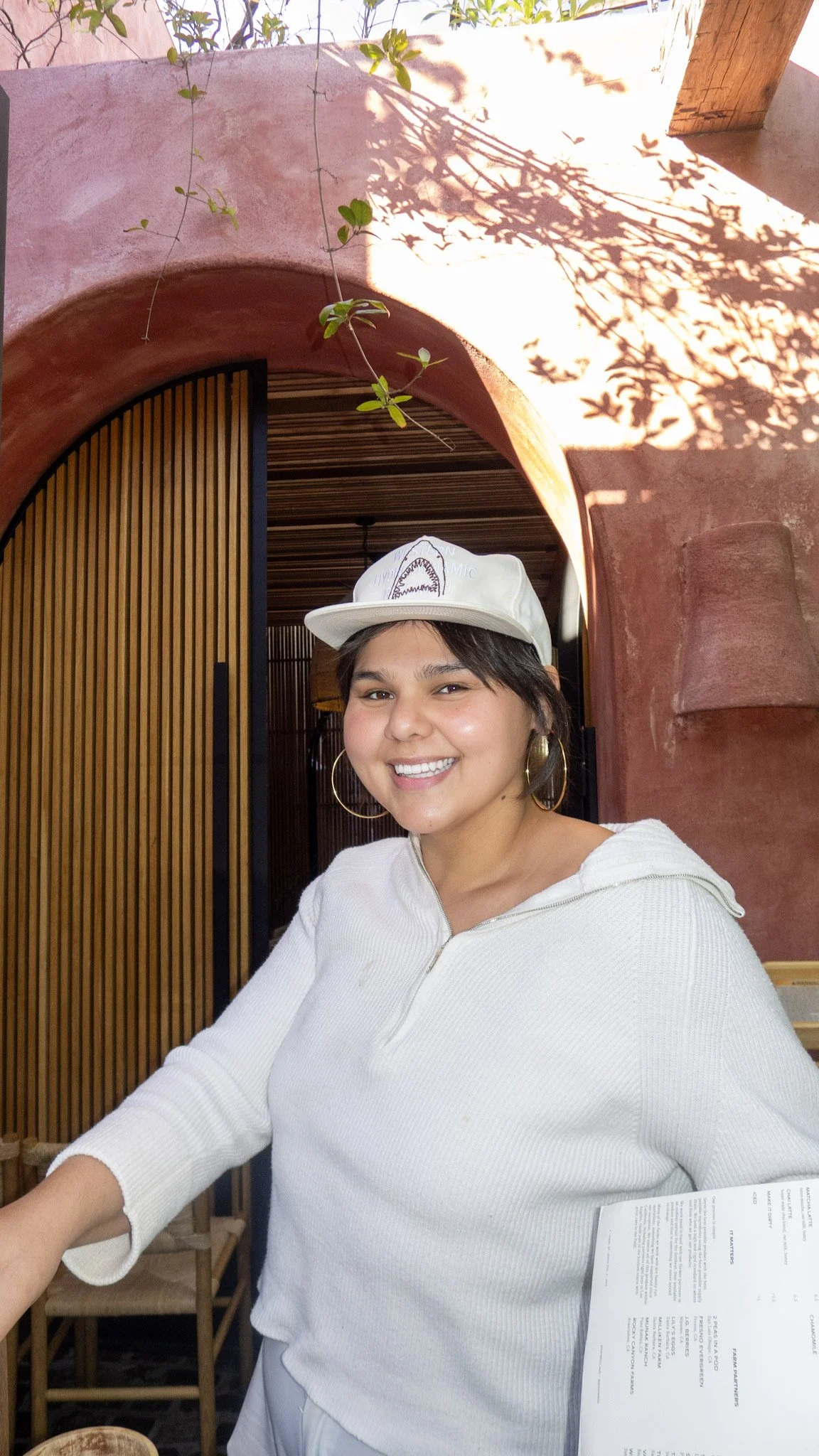 A smiling woman wearing a white cap and hoop earrings sitting inside a restaurant or cafe with wooden decor and sunlight casting shadows of leaves on the wall.