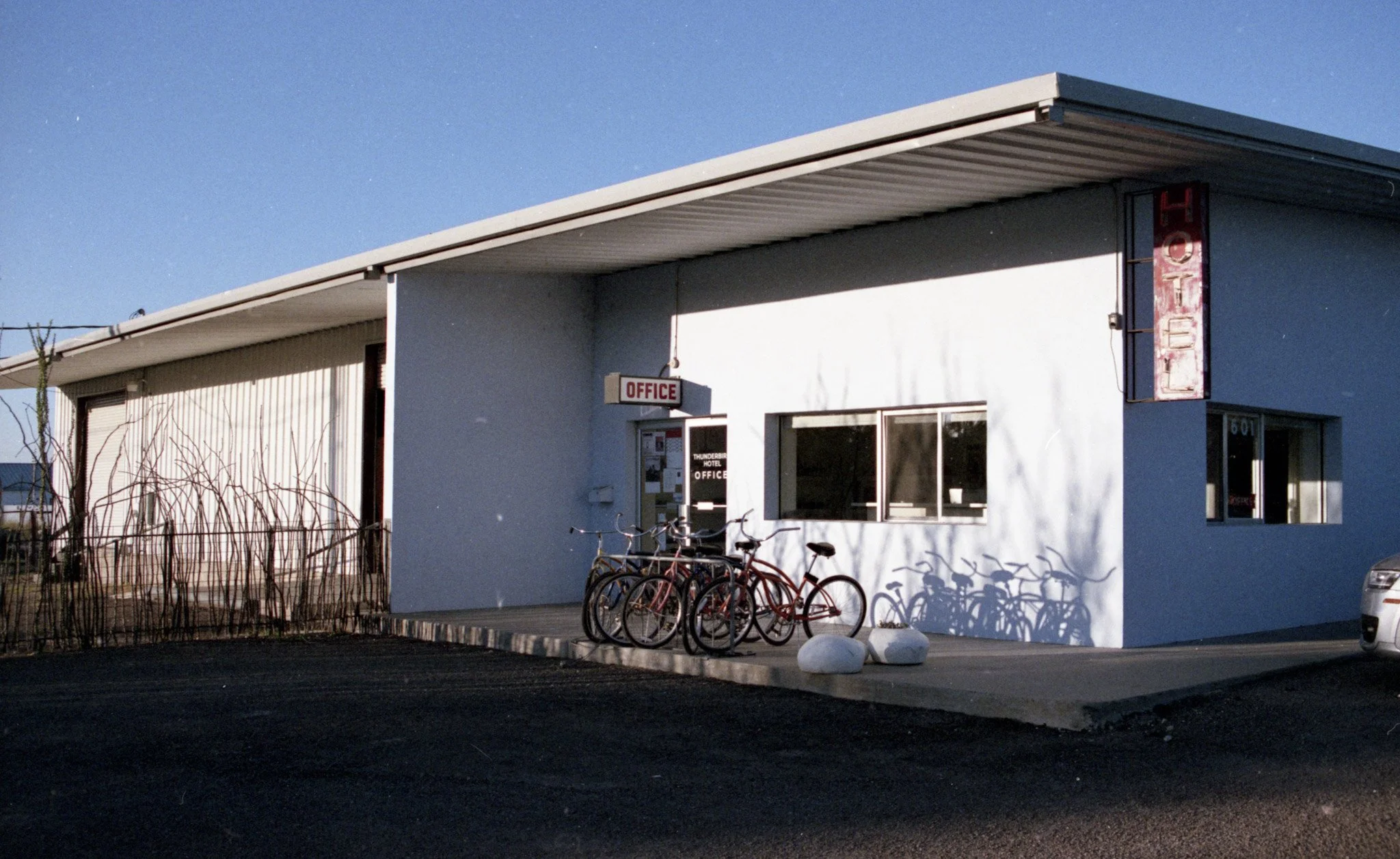 A small white building with a flat roof and an office sign near the entrance. Multiple bikes are parked in front, and there are small plants or bushes along the side. The sky is clear and blue, suggesting daytime.